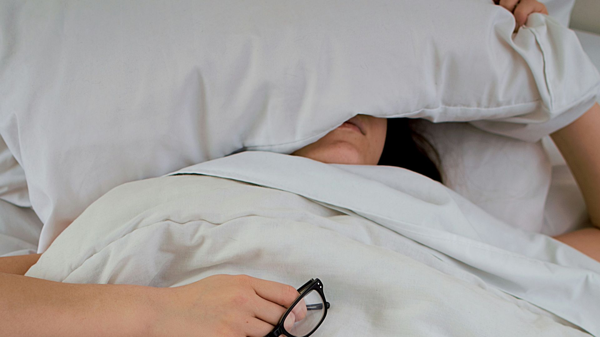 person lying on bed while covering face with pillow and holding eyeglasses