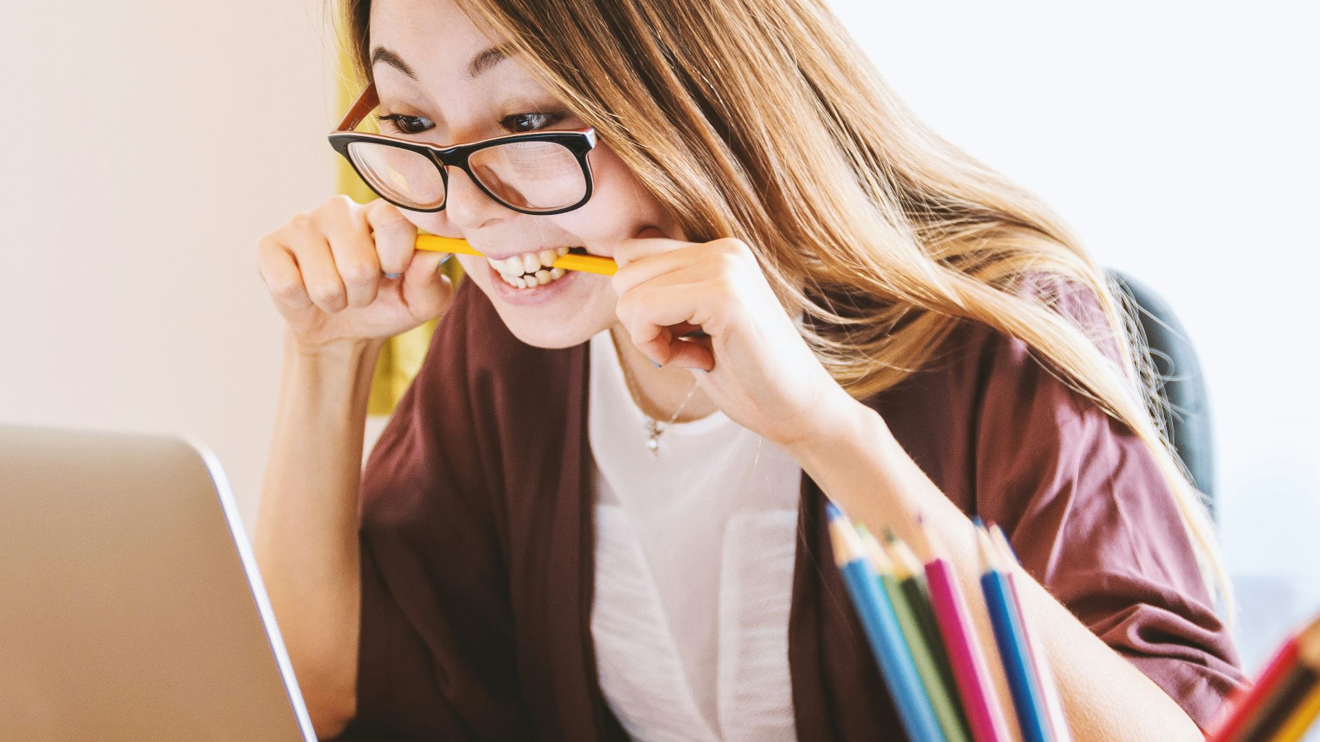 woman biting pencil while sitting on chair in front of computer during daytime