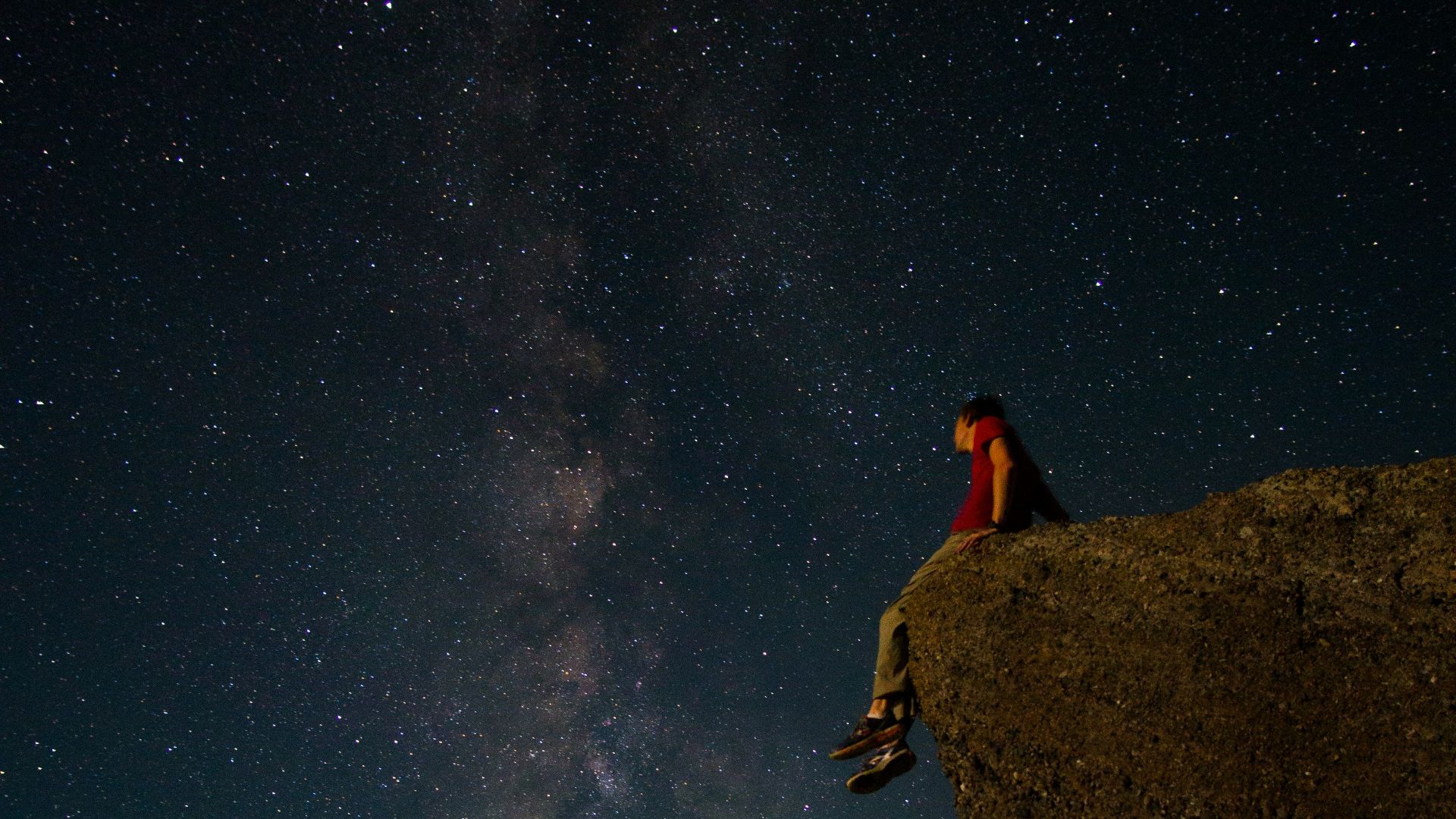 person sitting on edge of large rock under starry night sky