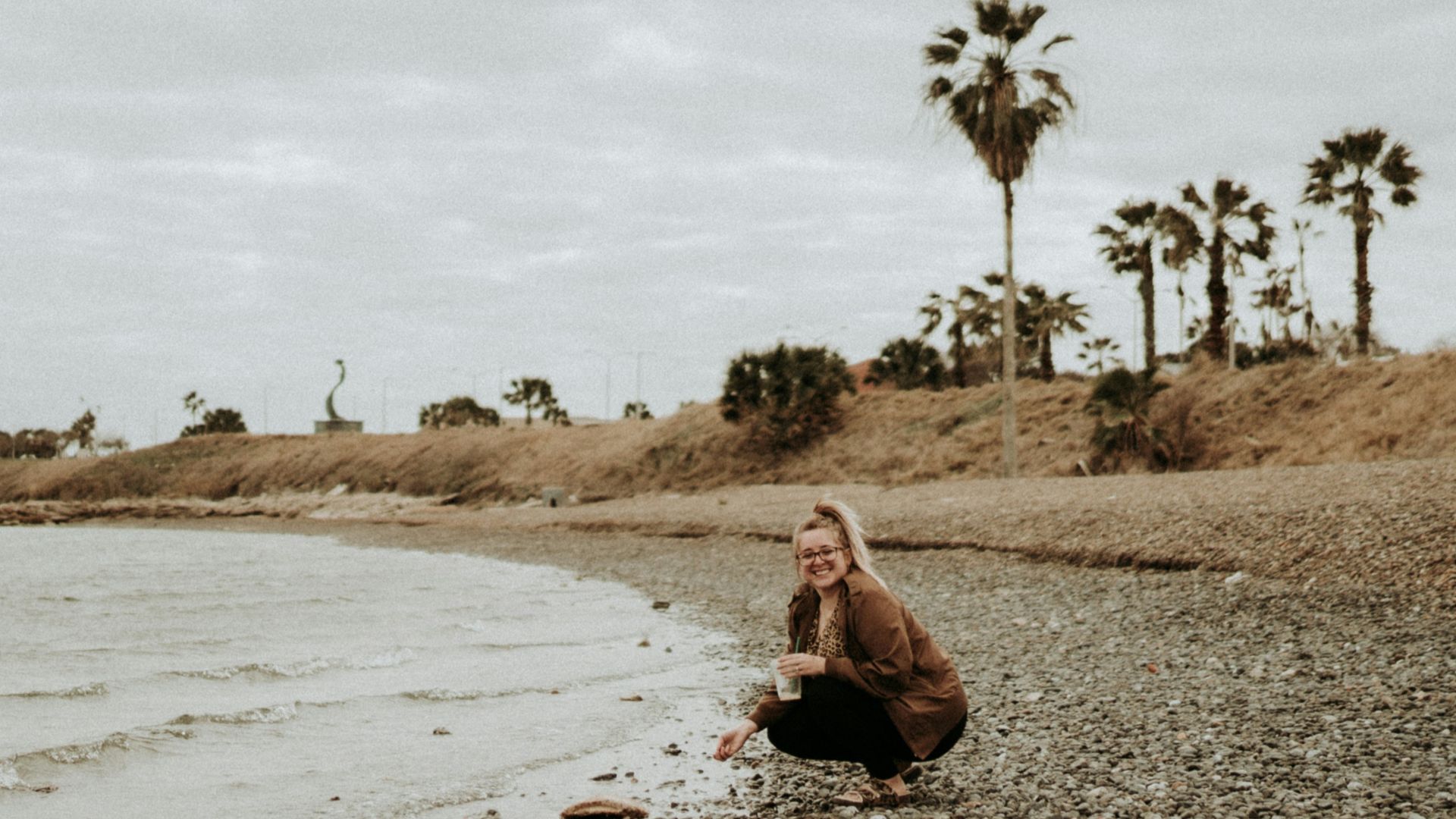 woman in brown jacket sitting on brown sand near body of water during daytime