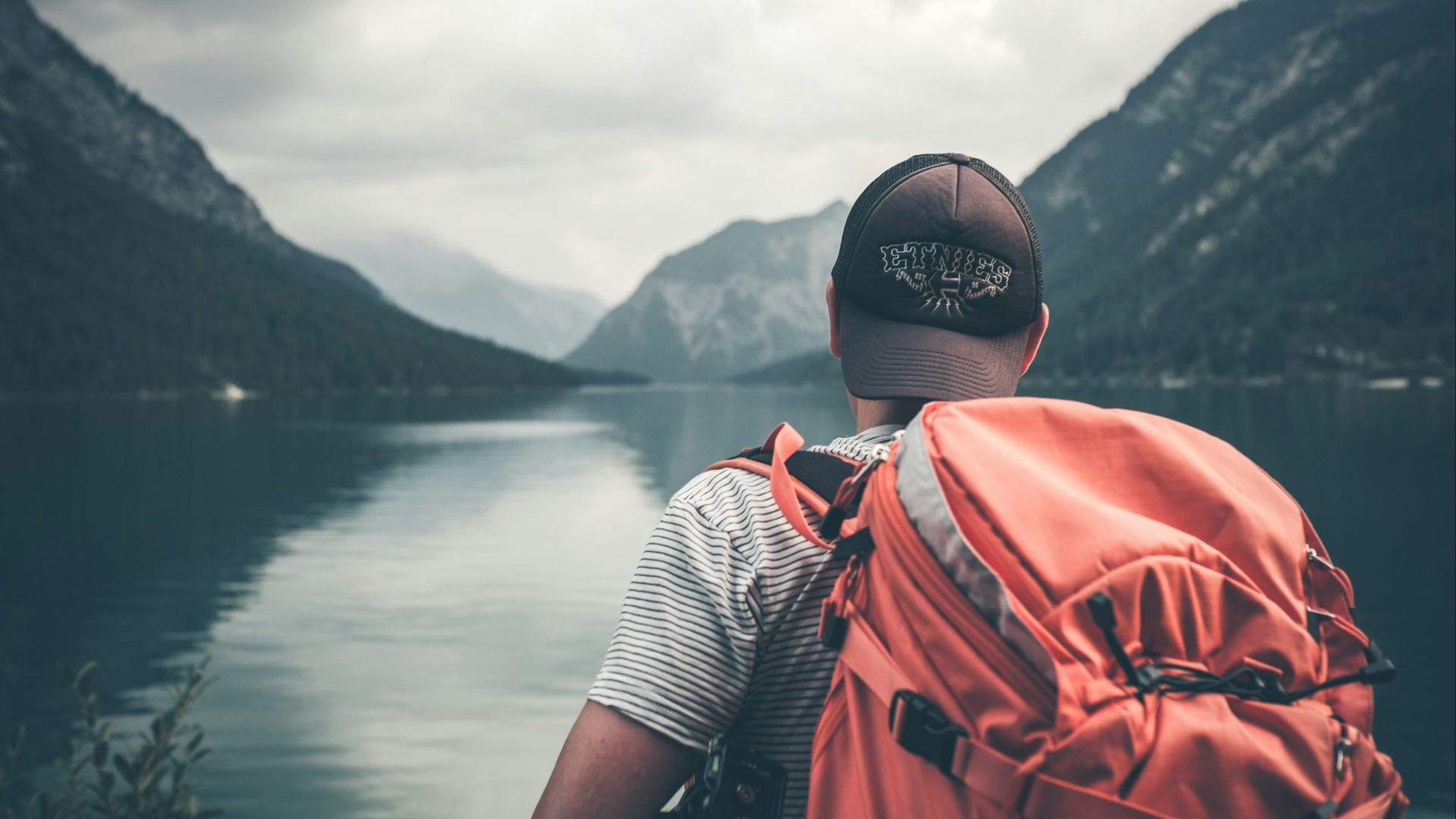 man with red hiking backpack facing body of water and mountains at daytime