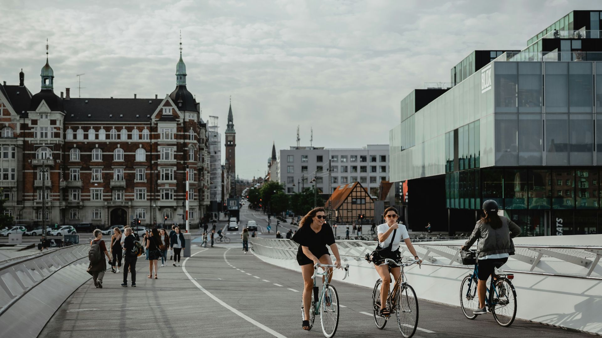 two people biking and other people walking on pathway near buildings under white and blue sky