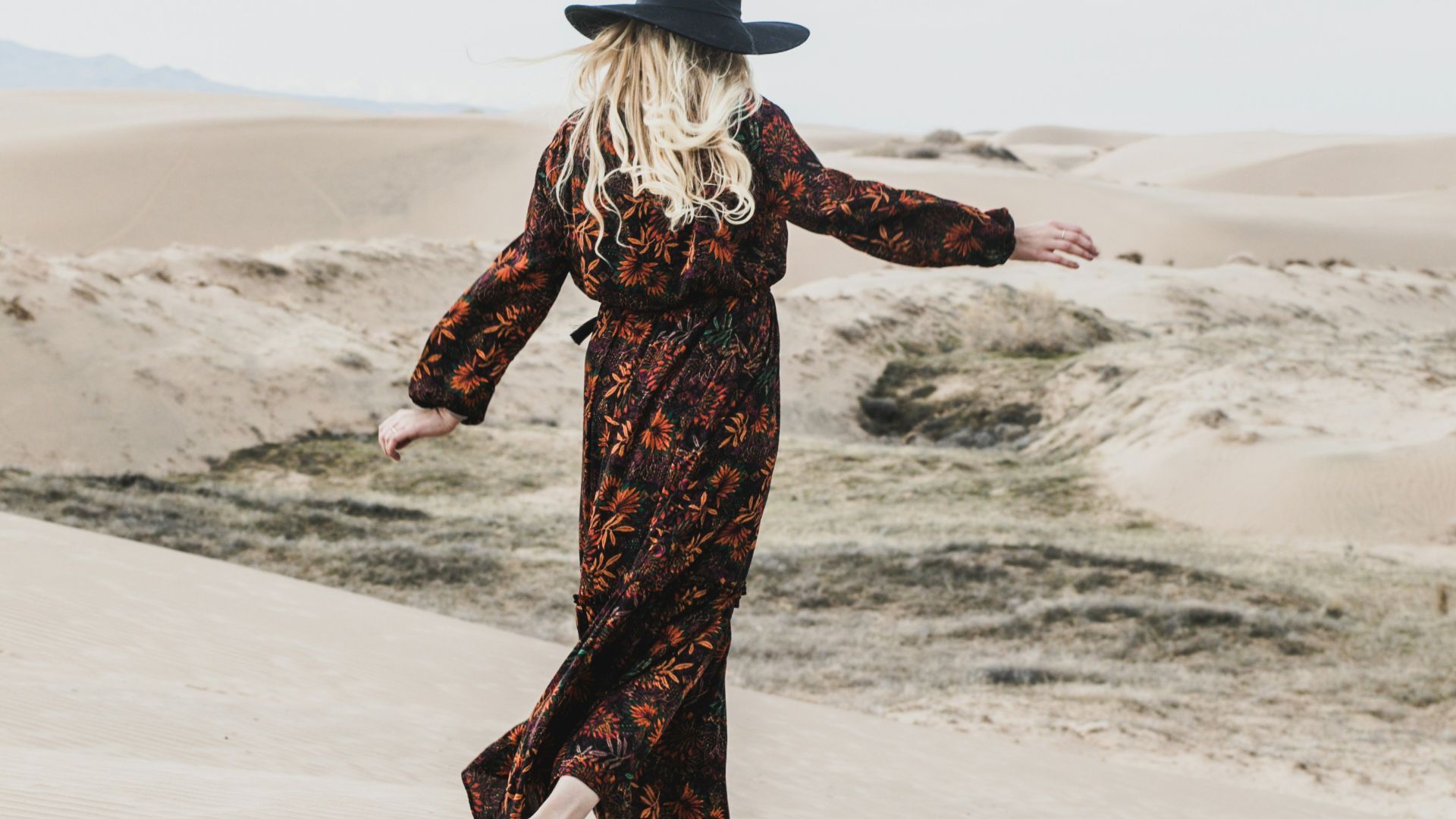 photo of person wearing brown and orange floral maxi dress walking barefooted along deserted land