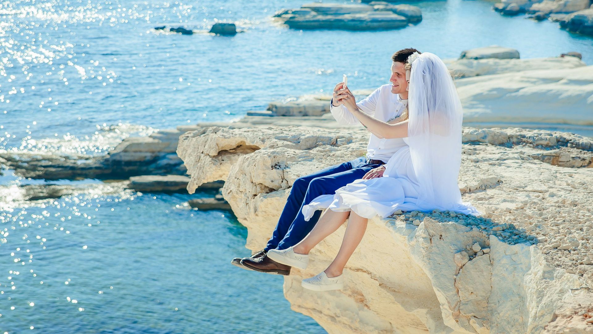 woman wearing wedding dress sitting beside man wearing white dress shirt on the stone tip facing the sea