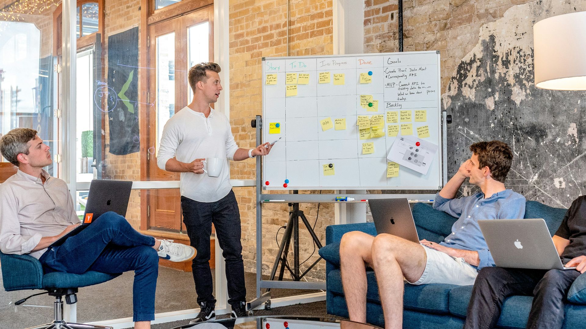 three men sitting while using laptops and watching man beside whiteboard