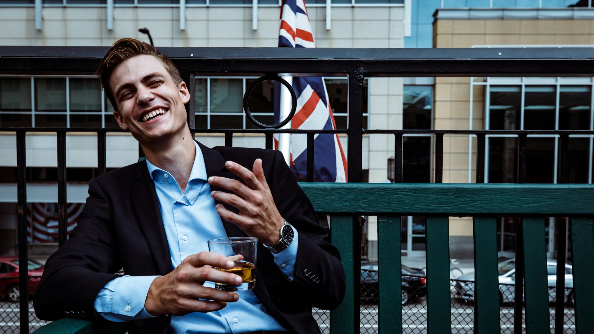 man smiling while sitting and holding whisky glass near concrete building