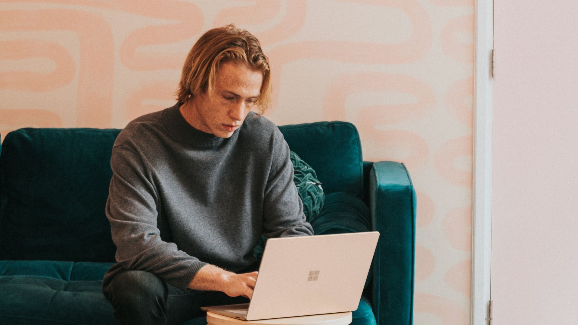 man in gray long sleeve shirt sitting on blue sofa working on a Microsoft Surface laptop