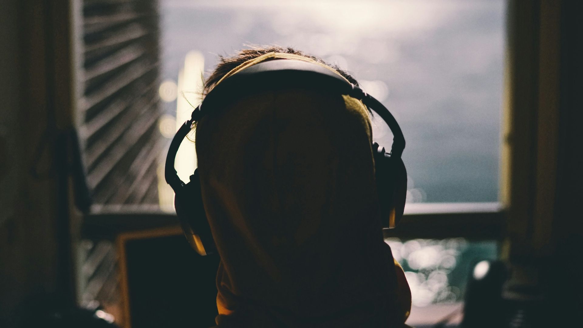 tilt-shift lens photography of person wearing headphones facing body of water through window of a dark room