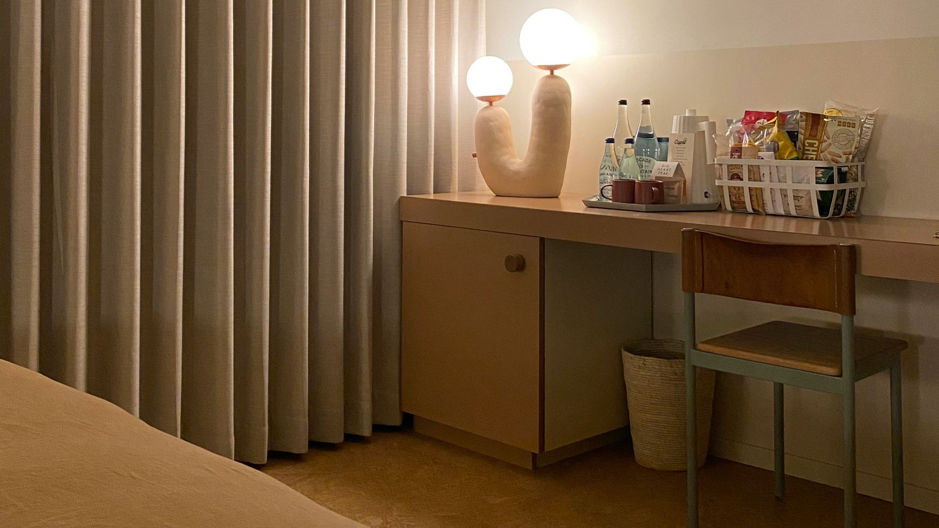 Hotel room desk with snacks, drinks, and lamp.