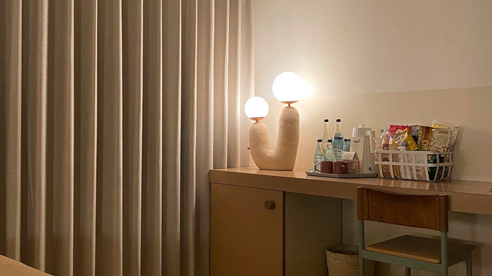 Hotel room desk with snacks, drinks, and lamp.