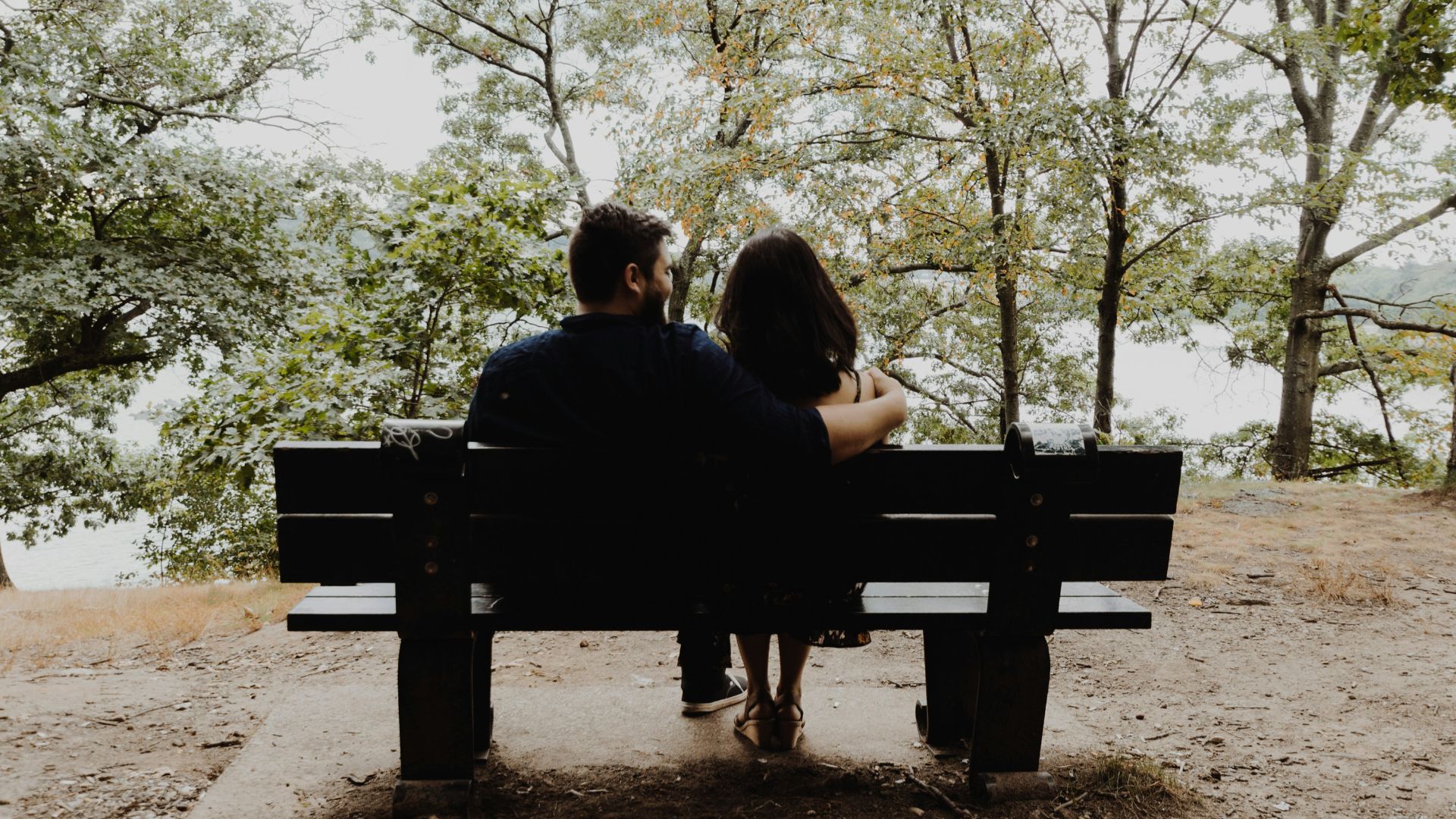 man looking to woman sitting on black wooden bench in front of tall trees during daytime