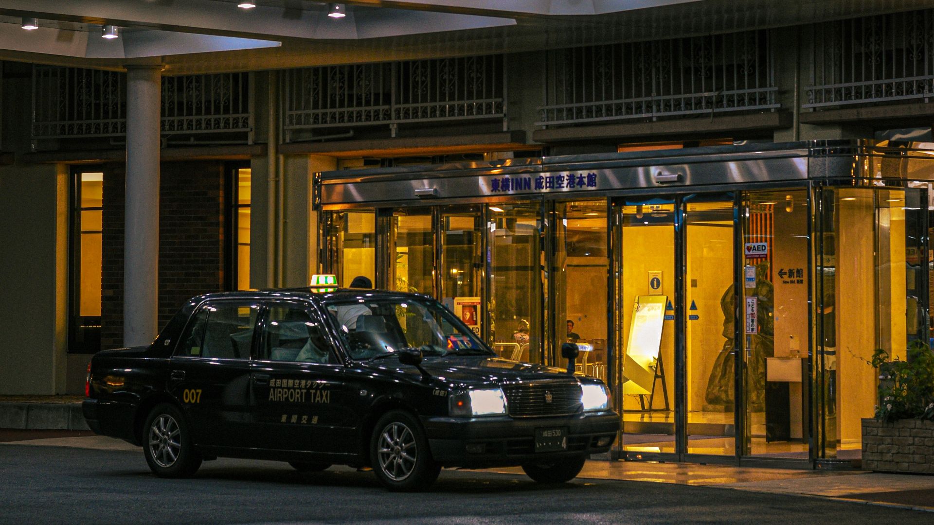 A black taxi waits outside a brightly lit hotel entrance.