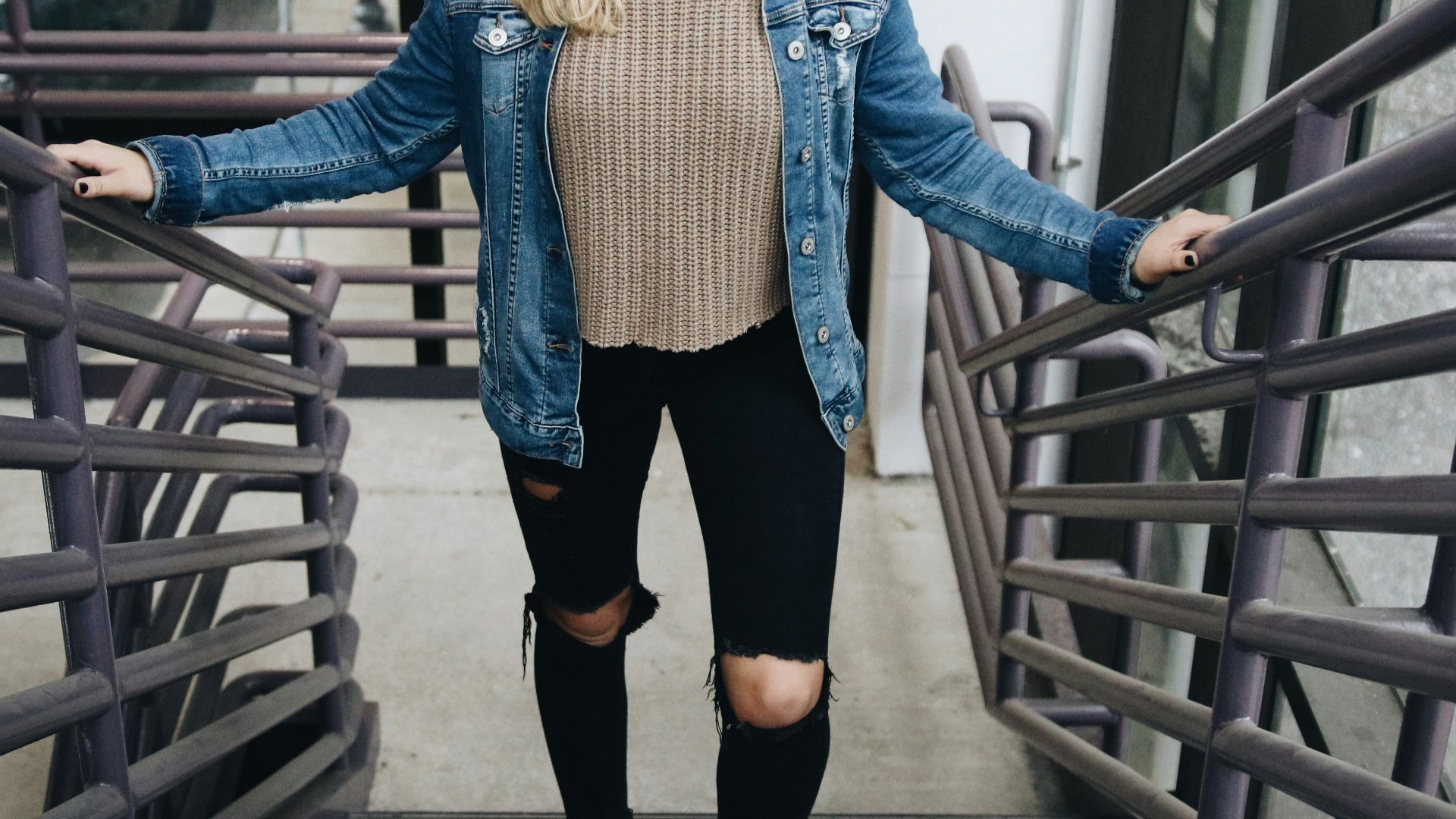 woman wearing blue denim jacket, brown sweater, black jeans, and hat standing in stair