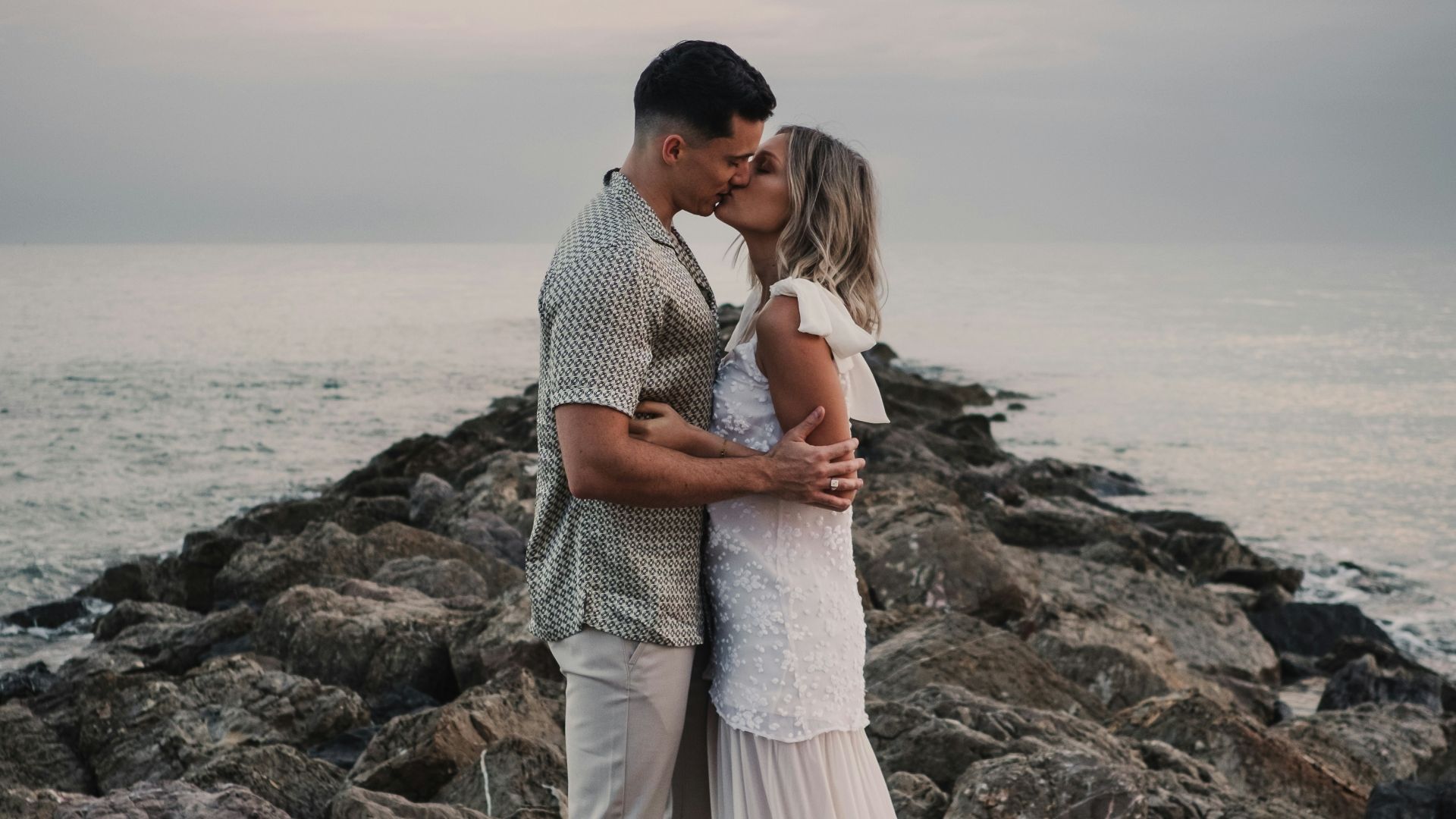 a bride and groom kissing on the rocks by the ocean