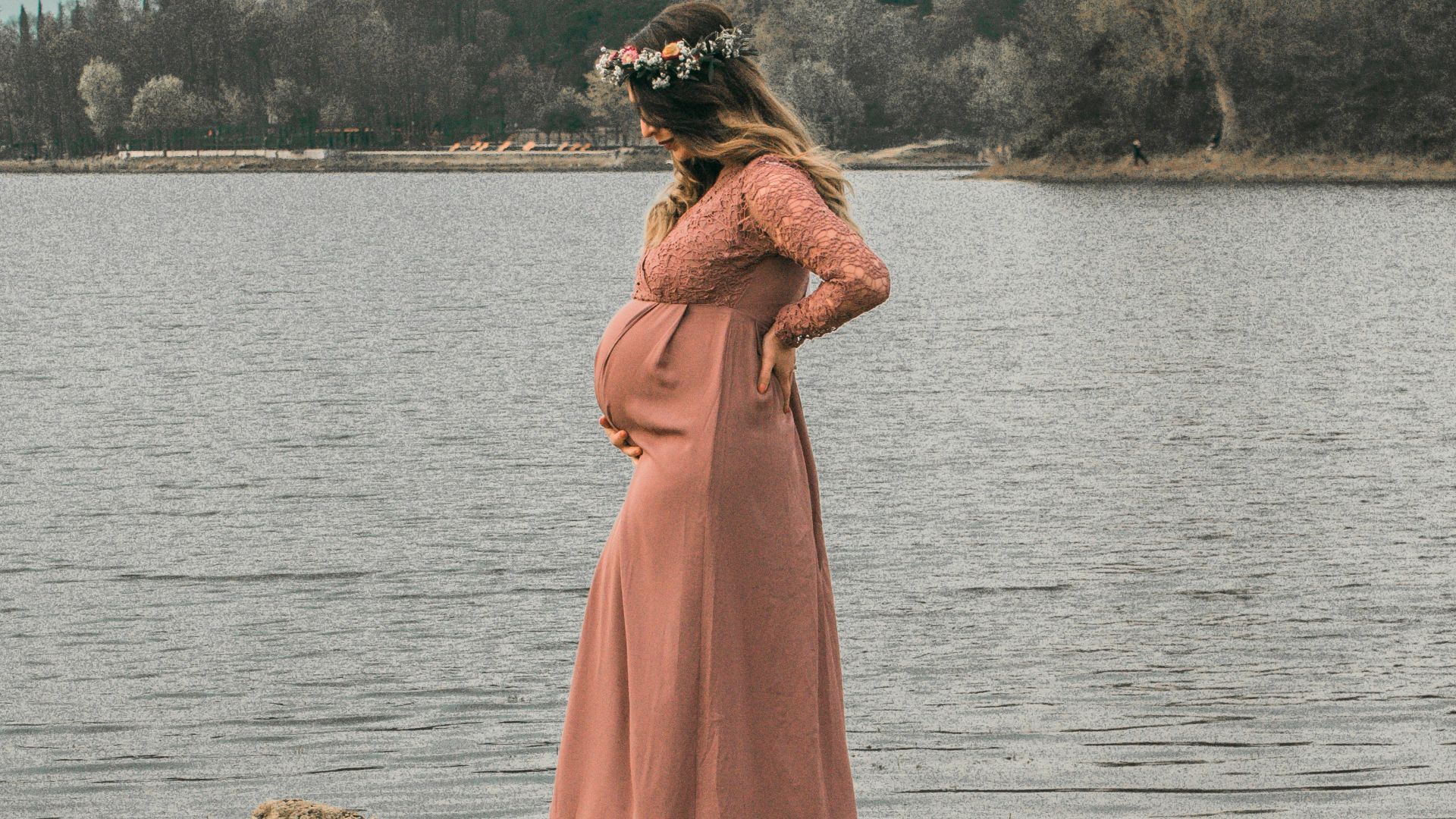 a pregnant woman in a pink dress standing on a rock by a lake