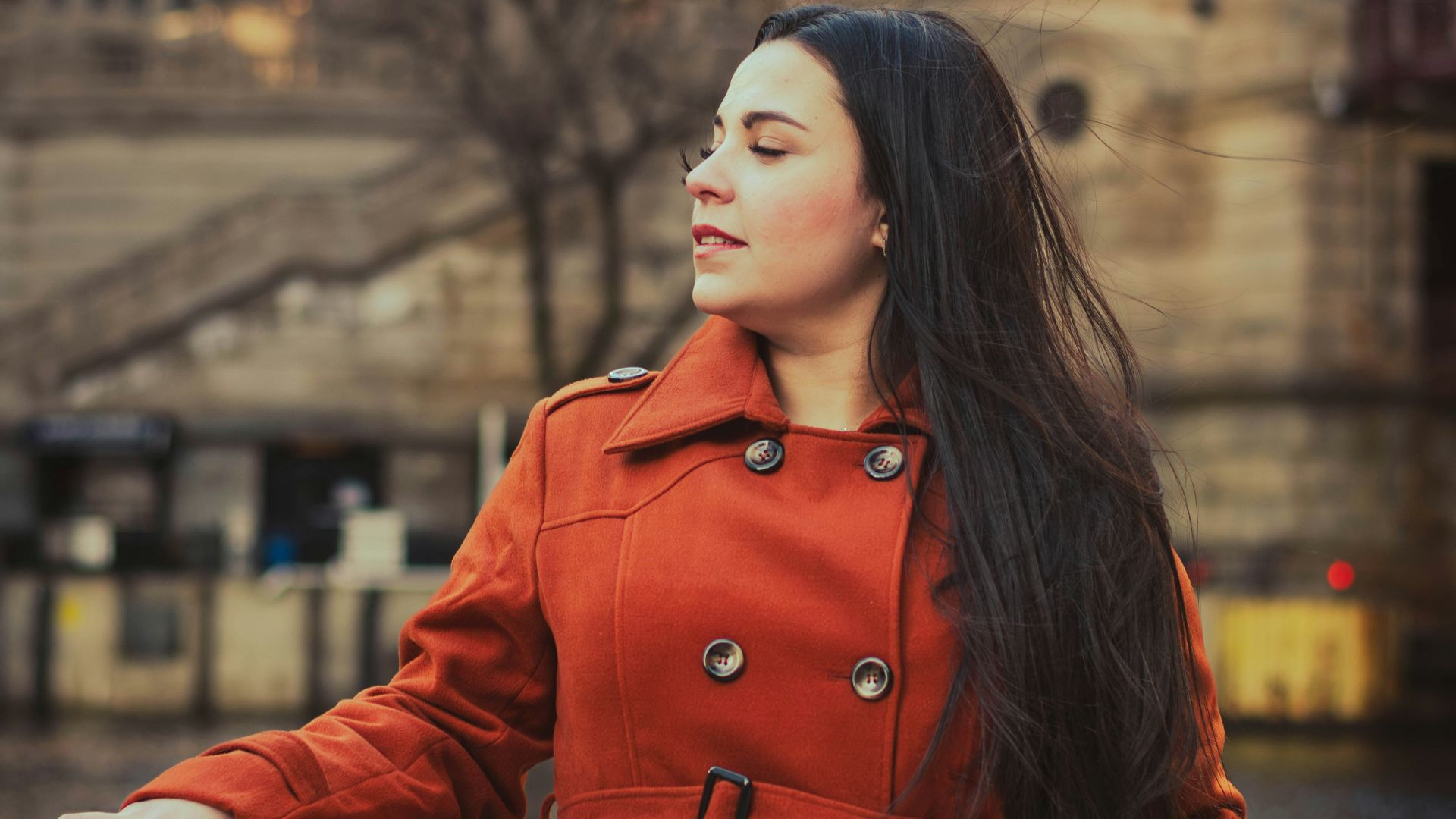 Woman in orange coat by river with city buildings