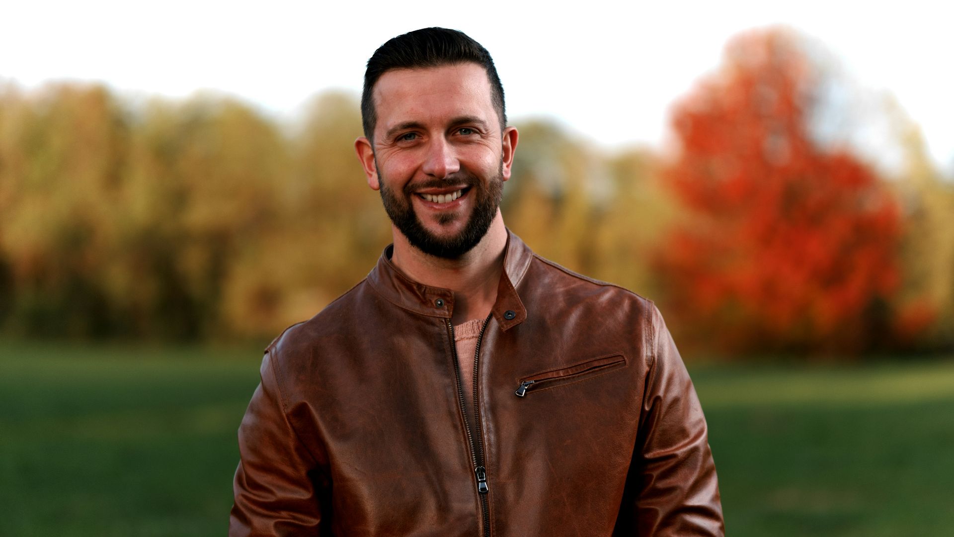 a man wearing a brown leather jacket standing in a field
