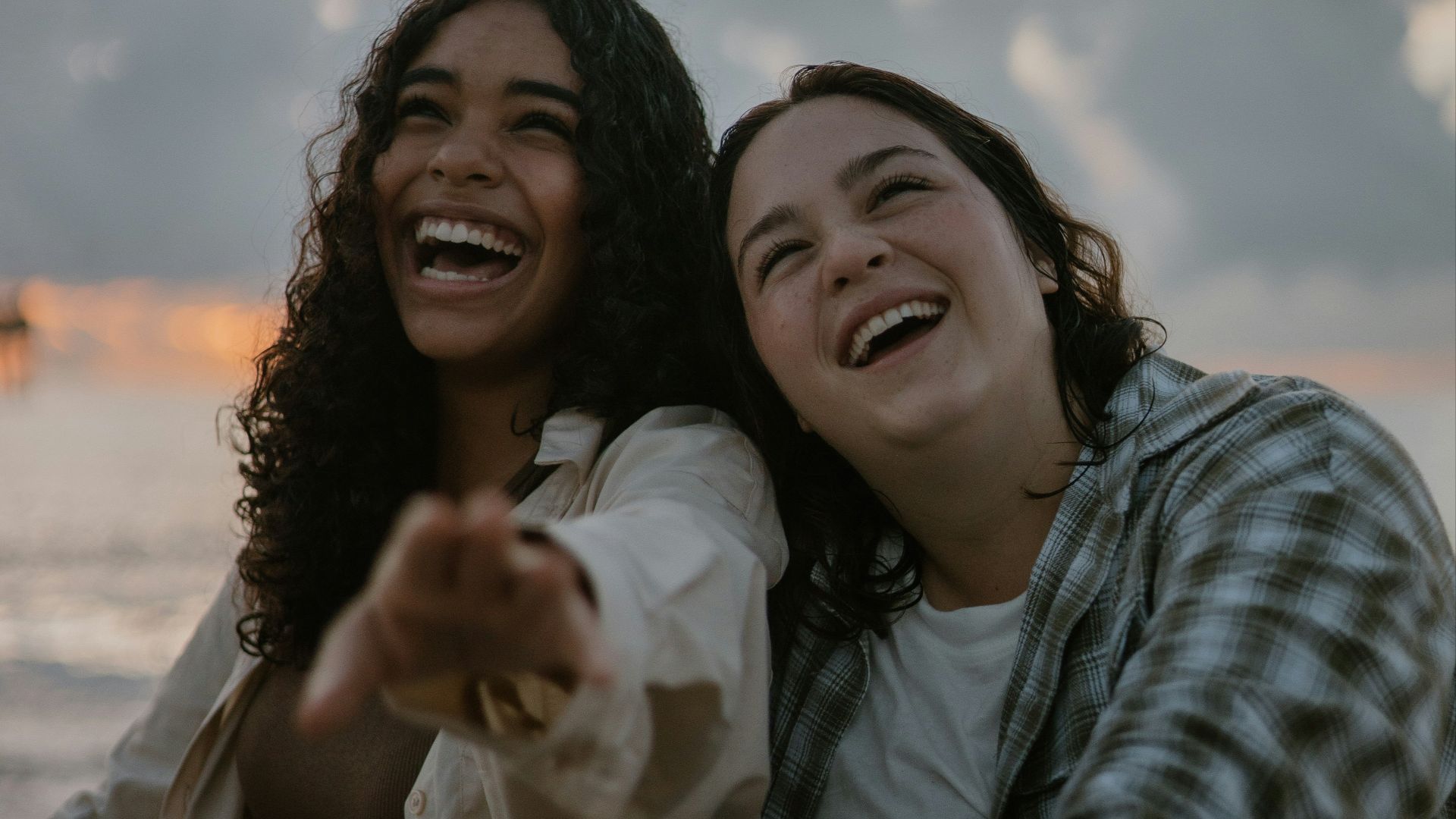 two women sitting on the beach pointing at the camera