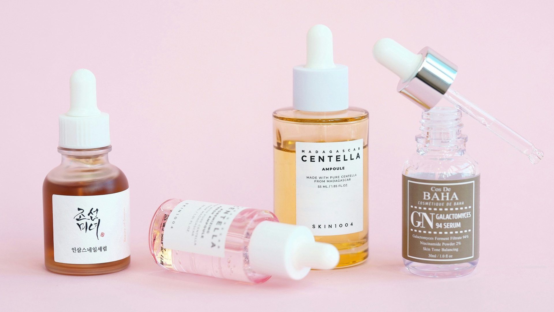 Skincare serums and dropper bottles sit on pink.