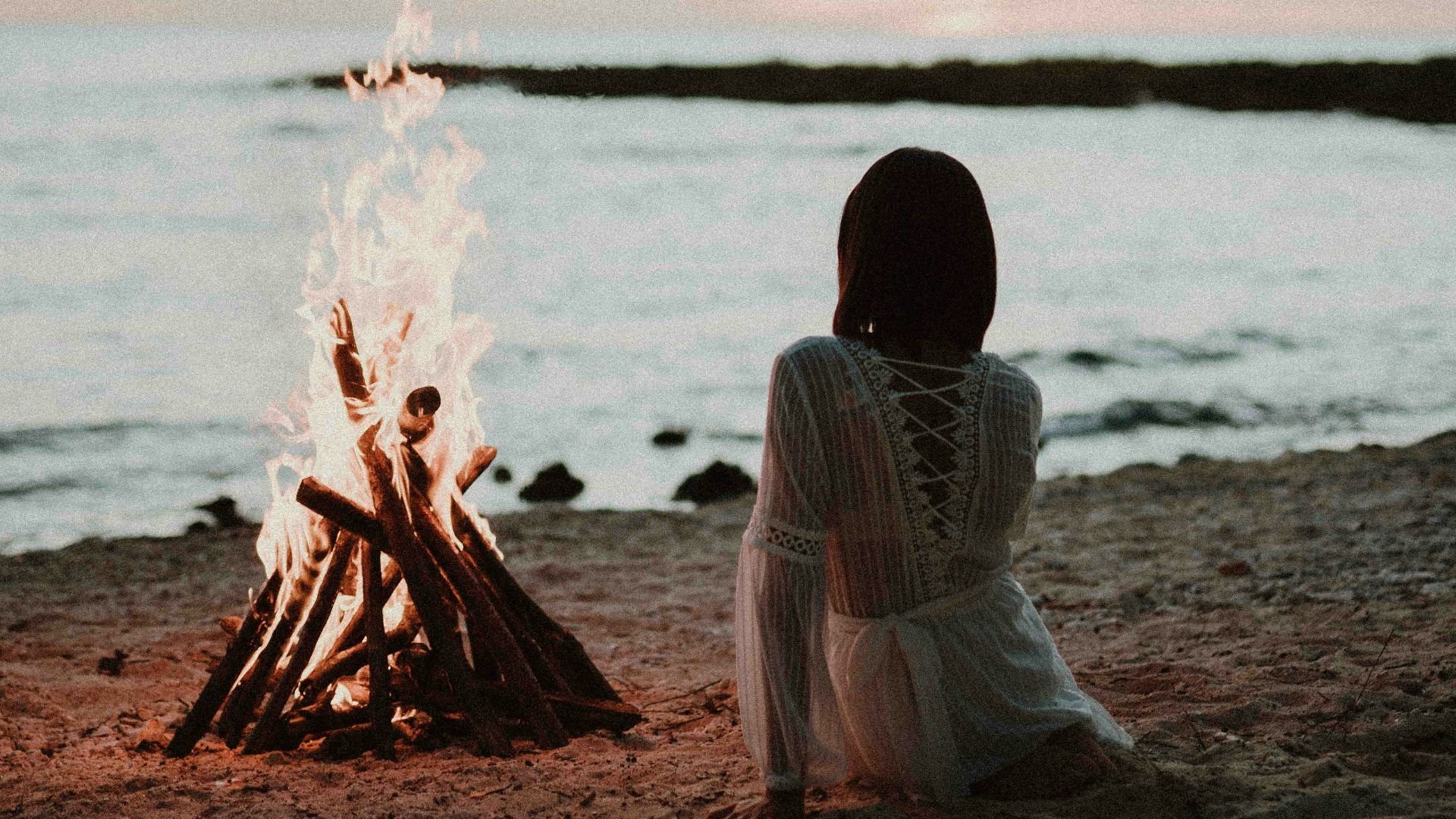 a woman sitting in front of a fire on a beach