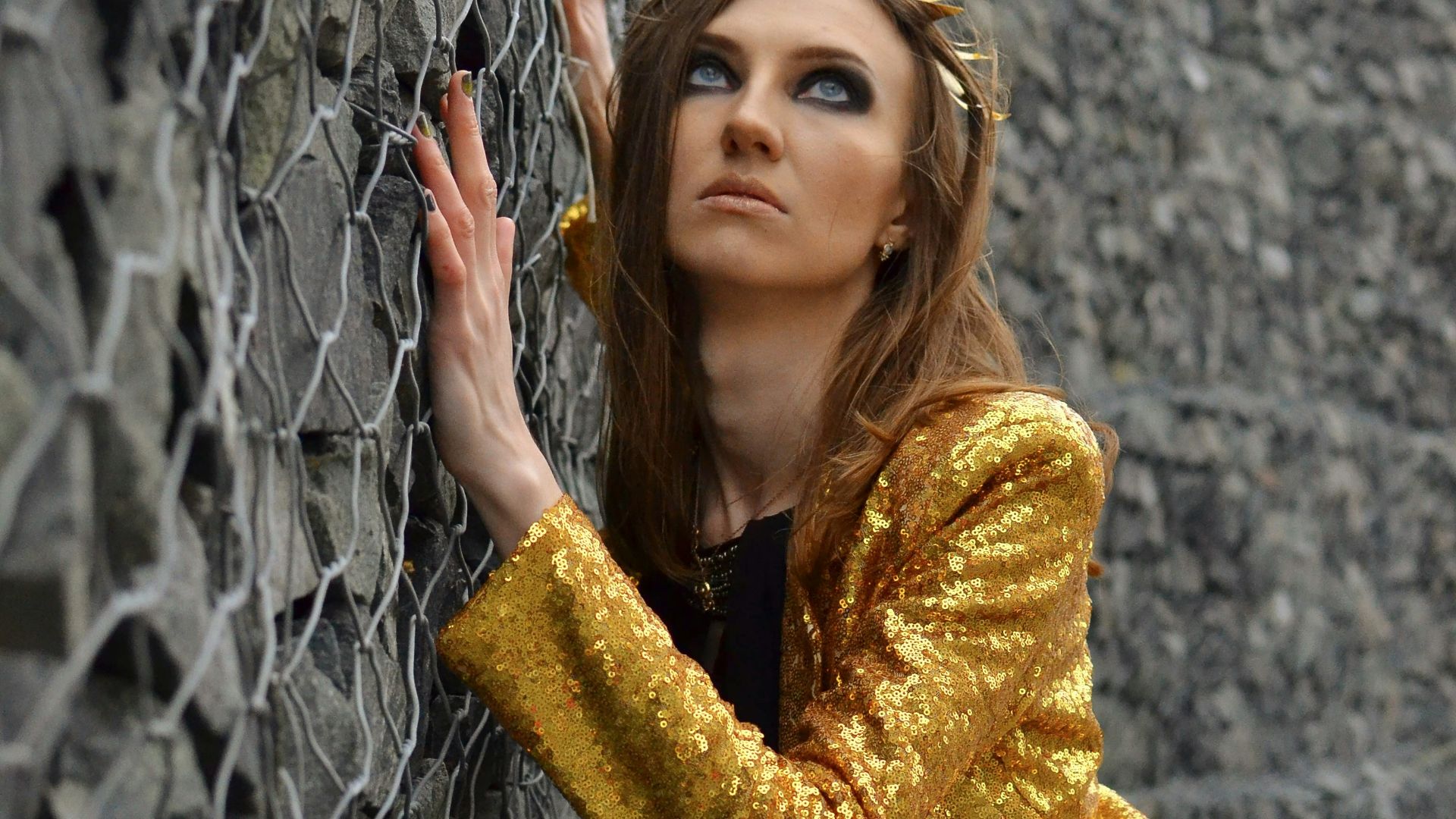 Woman with golden crown and jacket leans on wall.