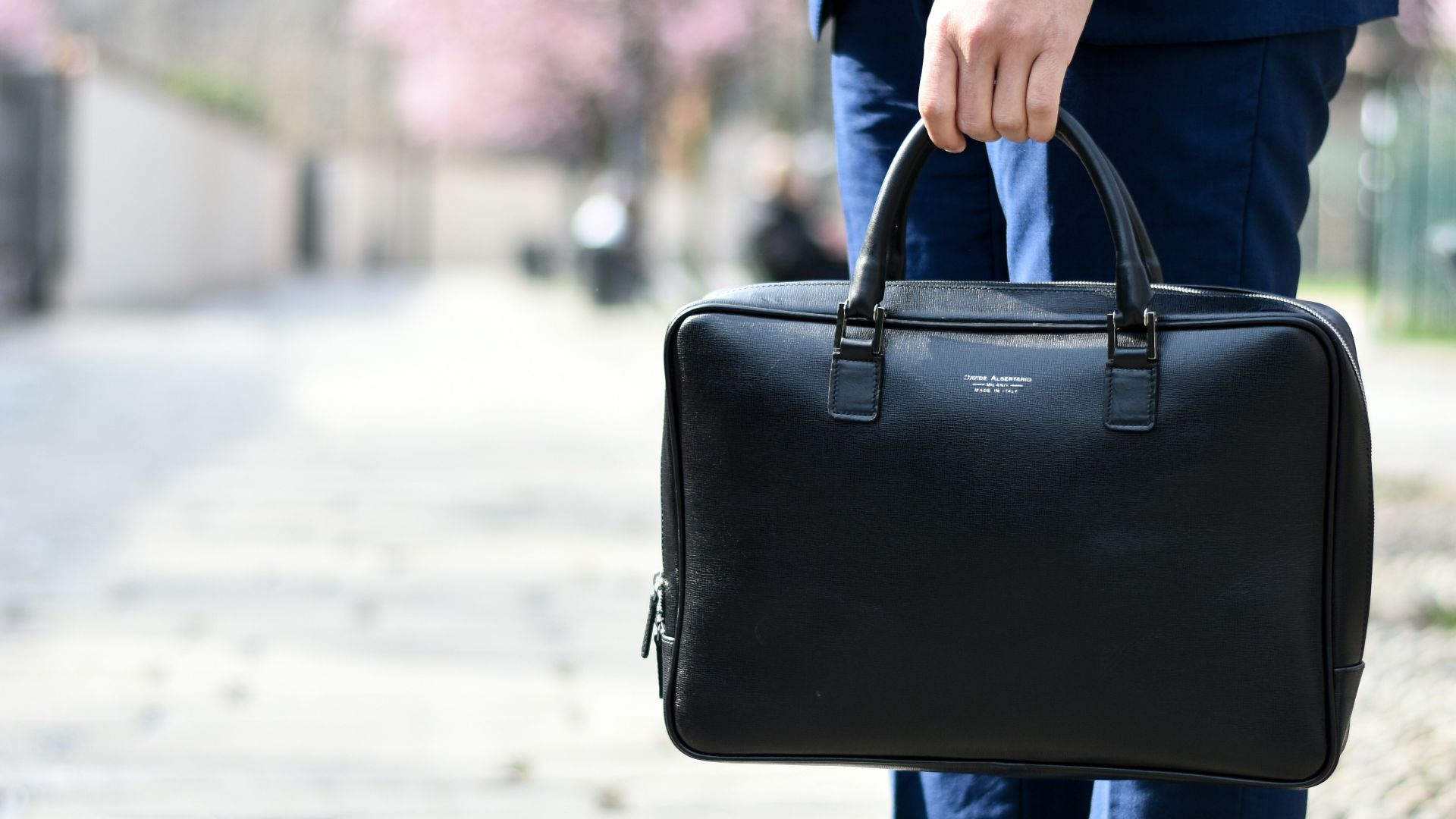 man in blue suit standing on street side while holding bag during daytime