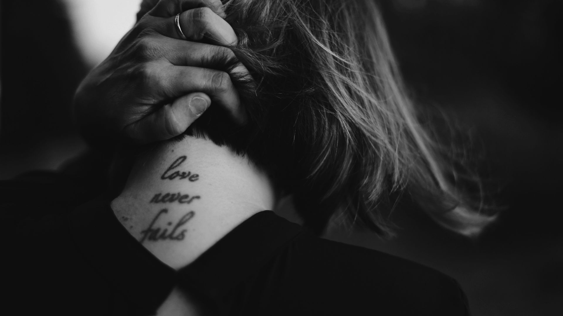 grayscale photo of woman holding her neck while showing Love never Fails tattoo at back