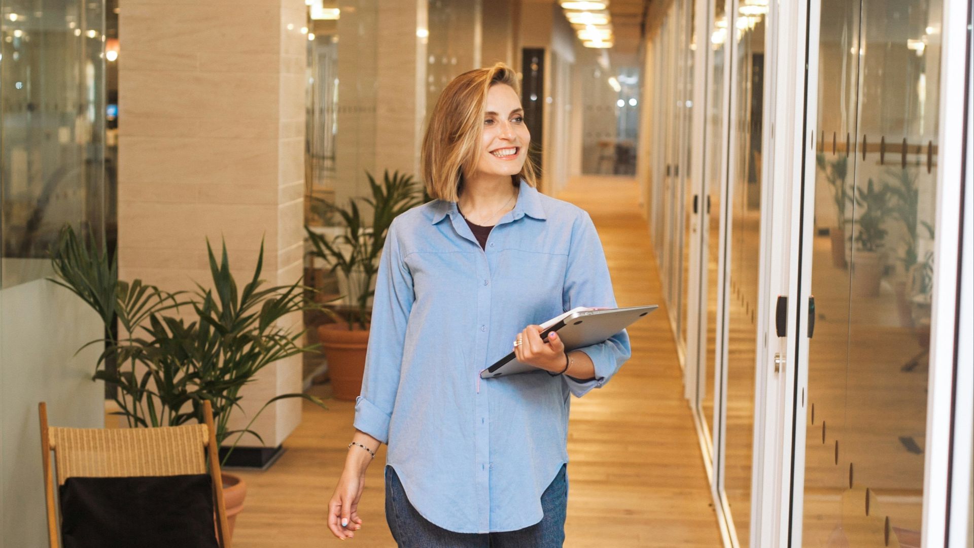 woman in blue dress shirt and blue denim jeans standing beside brown wooden chair