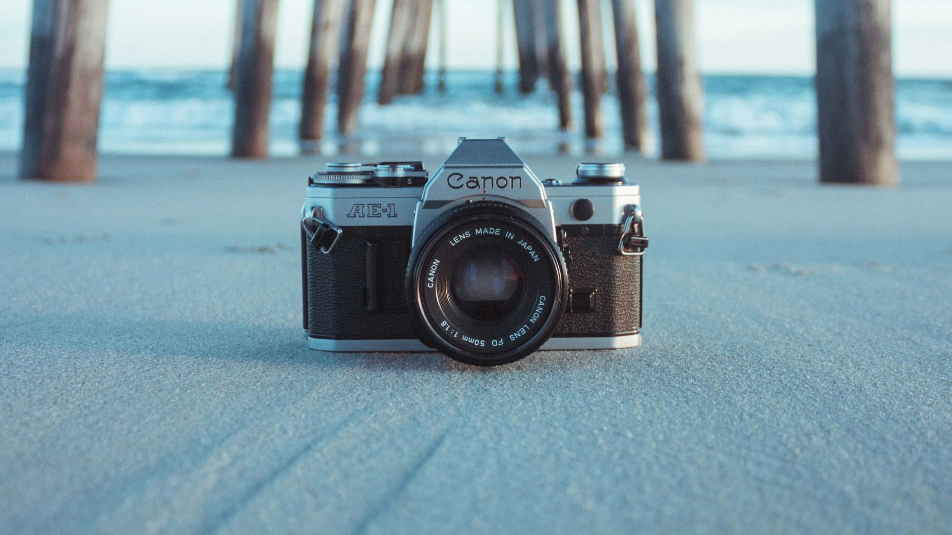 black and gray Canon AE-1 camera on gray sand under brown dock near body of water at daytime
