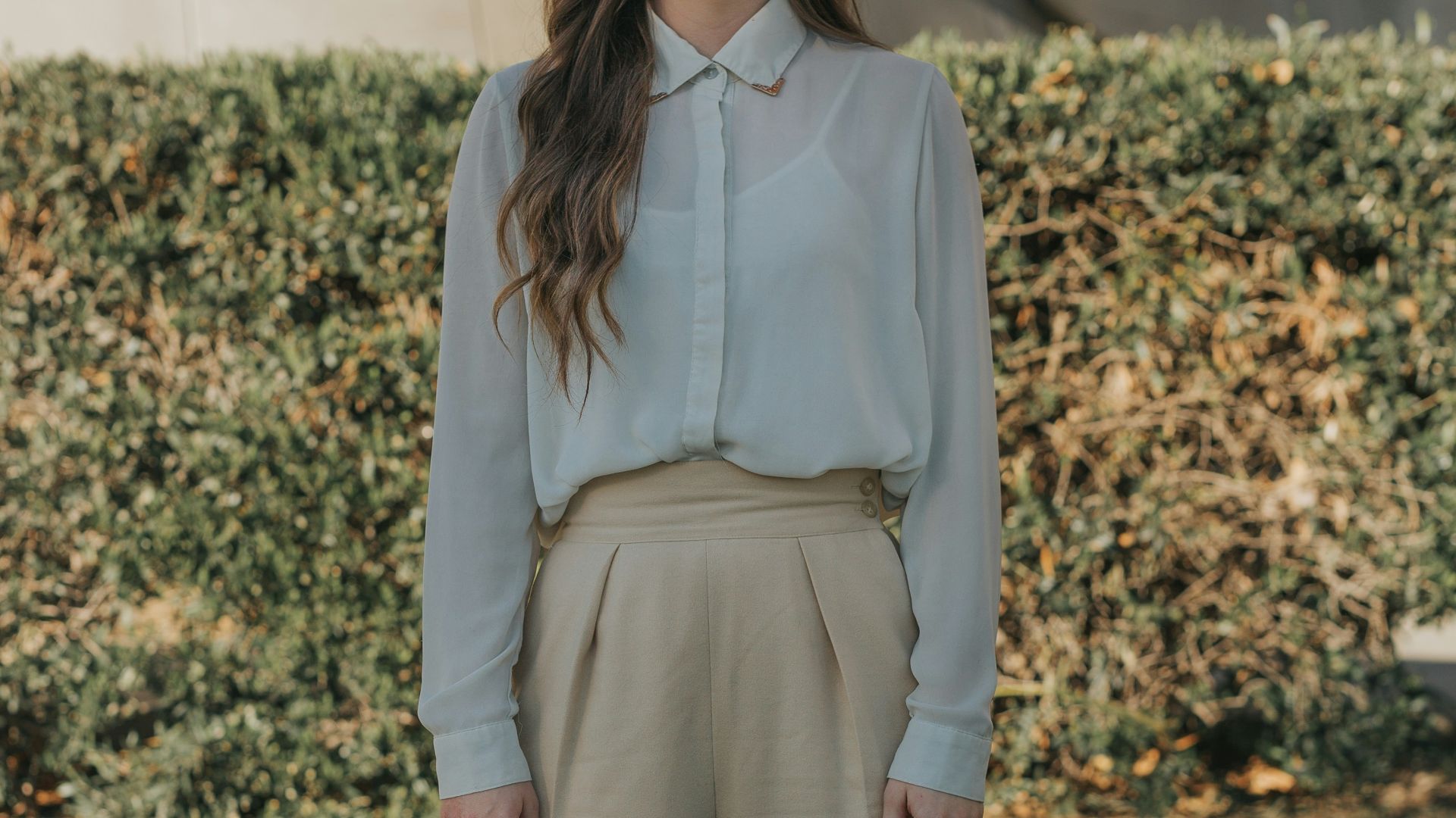 woman wearing gray button-up long-sleeved shirt and brown pants standing in front of bushes