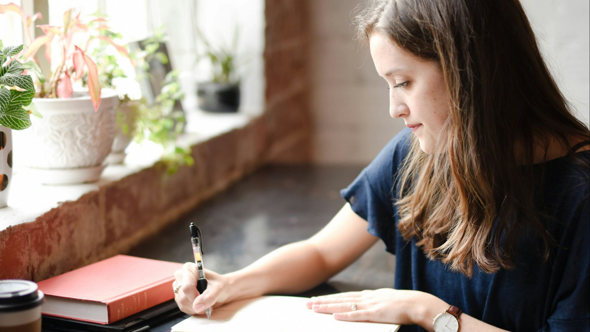 woman sitting in front of black table writing on white book near window