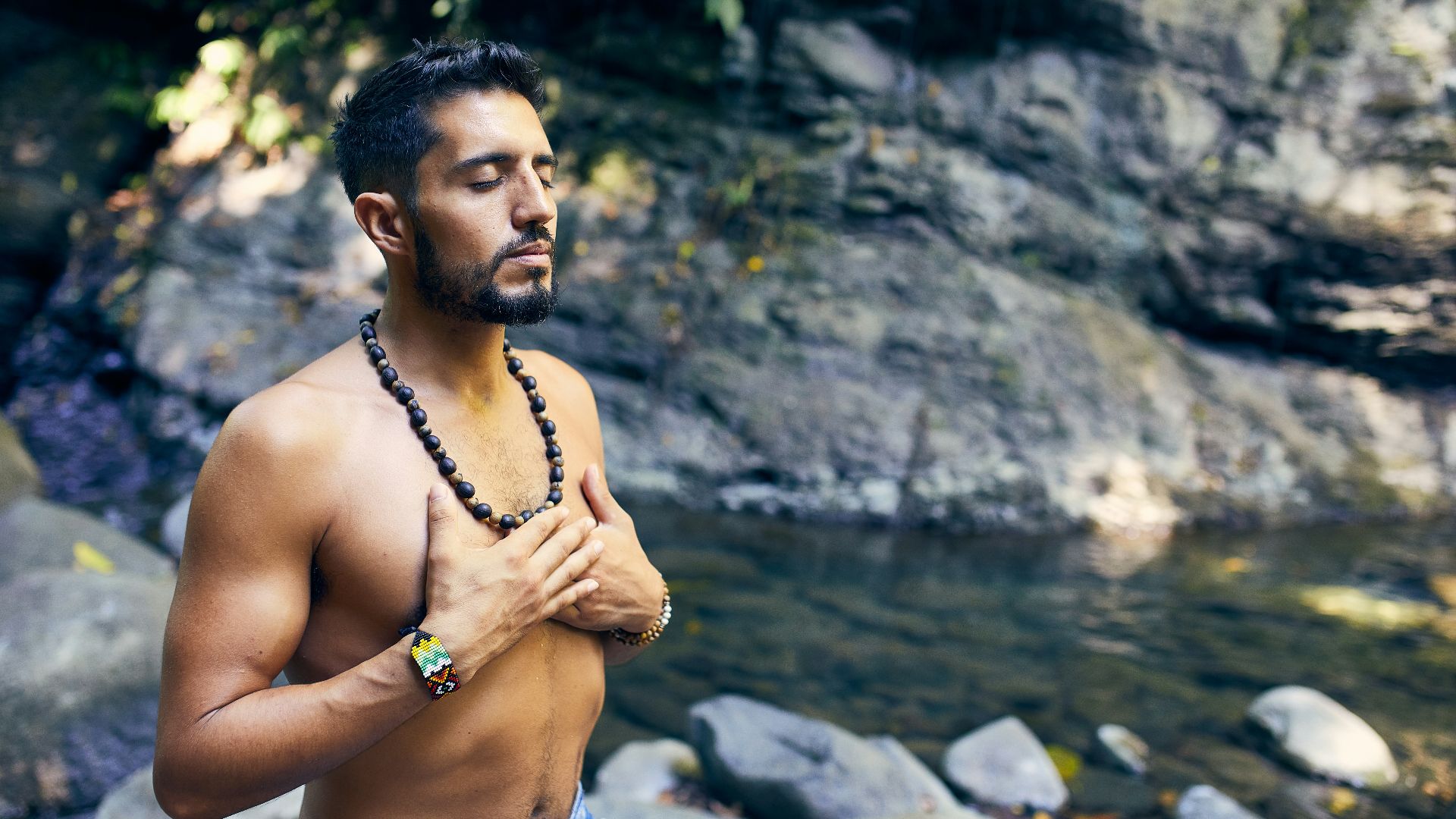 topless man wearing black beaded necklace and blue denim shorts standing on rocky shore during daytime