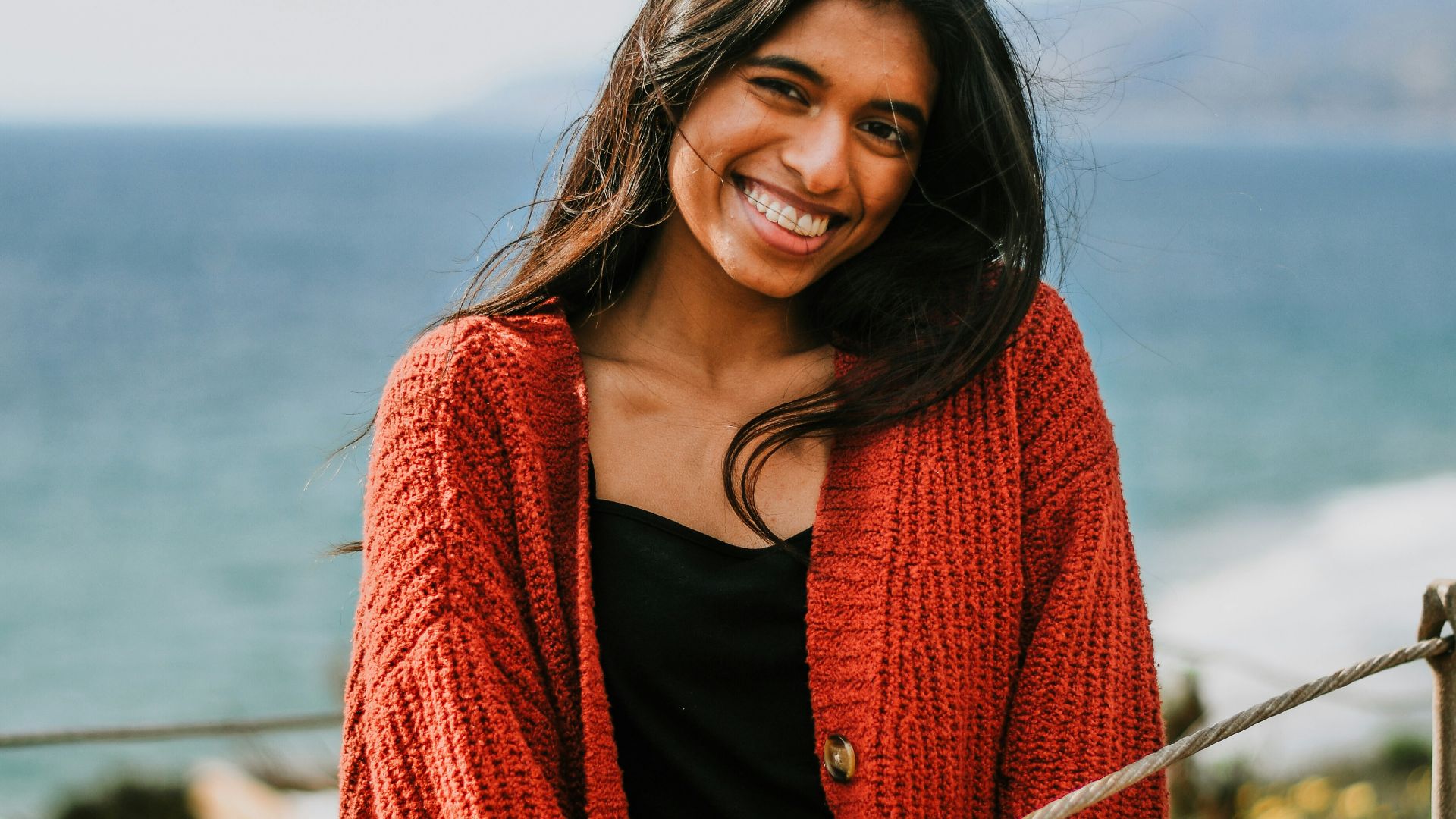 a smiling woman in a red cardigan by the ocean