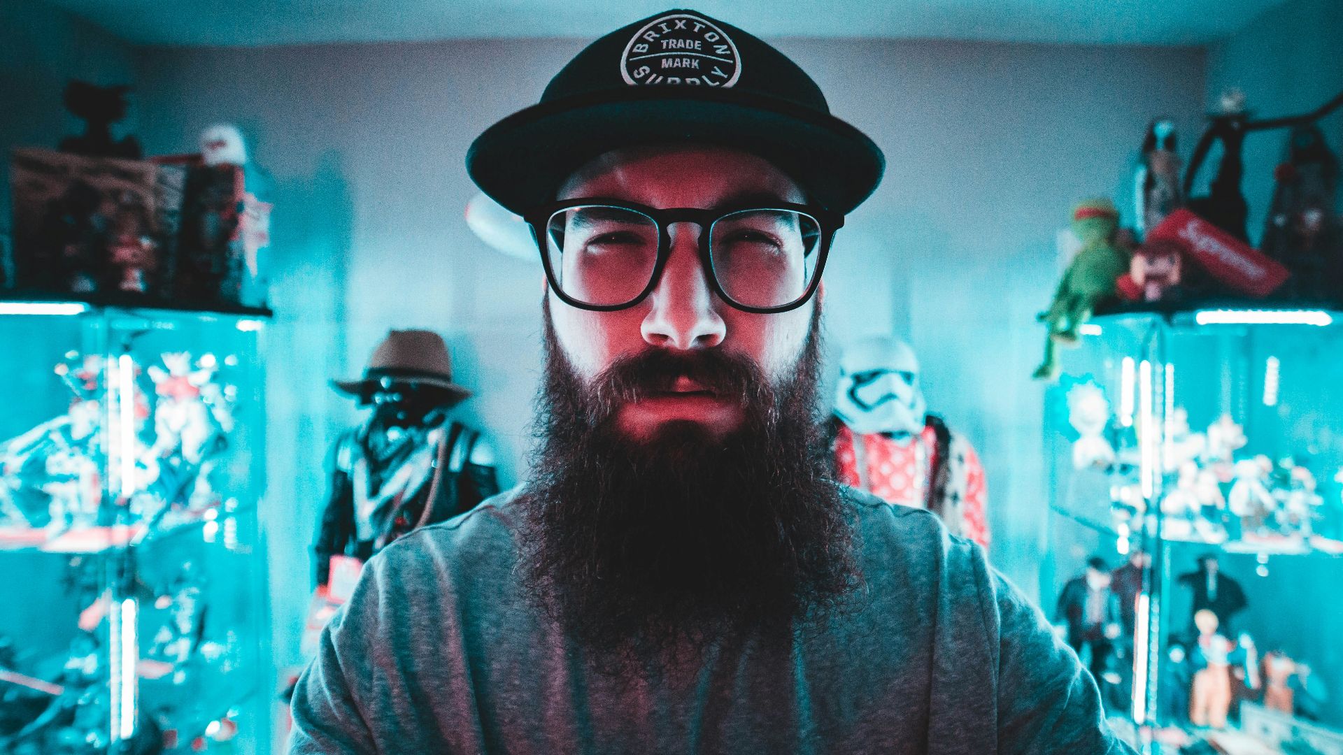 man wearing gray long-sleeved shirt, black and white hat, and eyeglasses inside green room