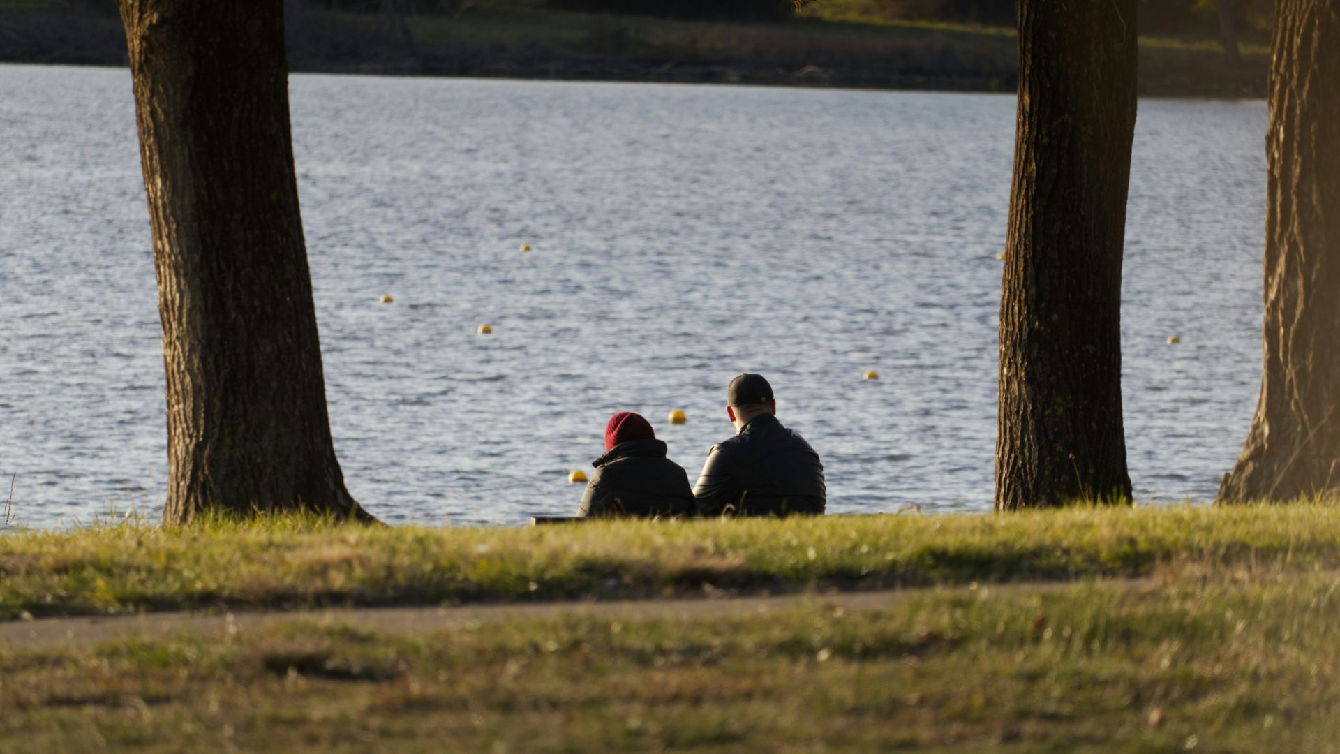 Two people sit by a calm lake at sunset