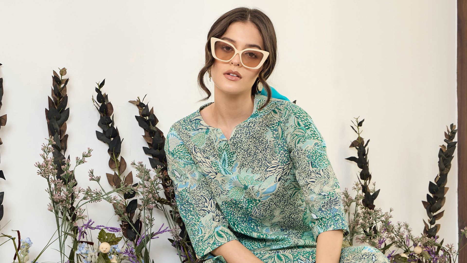 Woman in patterned outfit and sunglasses sits near plants.