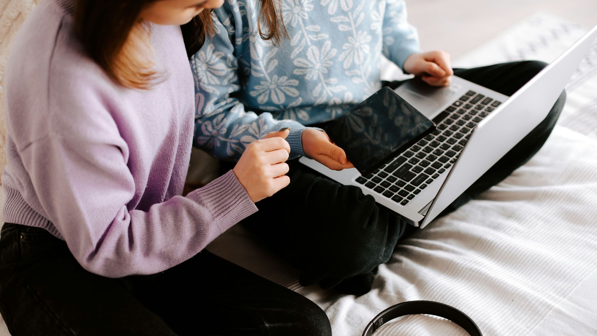 two girls sitting on a bed with a laptop and headphones