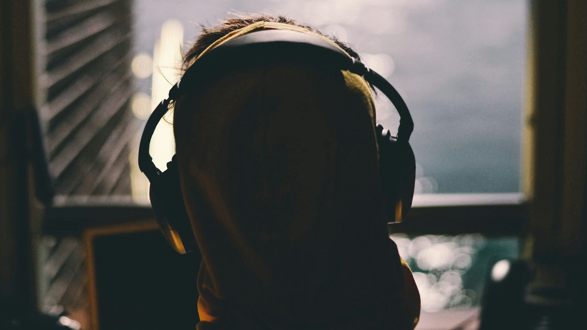 tilt-shift lens photography of person wearing headphones facing body of water through window of a dark room