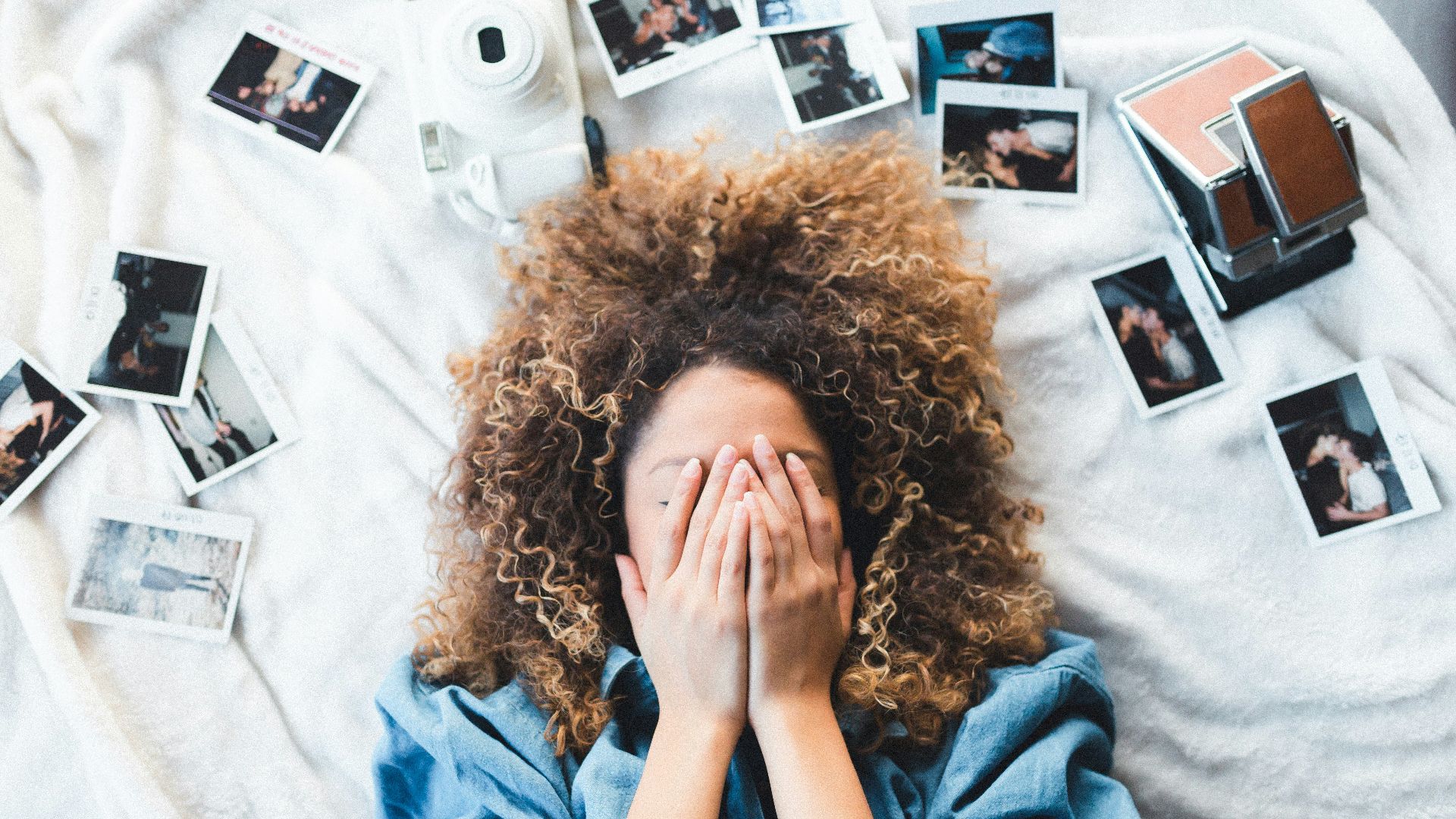 woman lying on bed covering her face surrounded by photos and white camera
