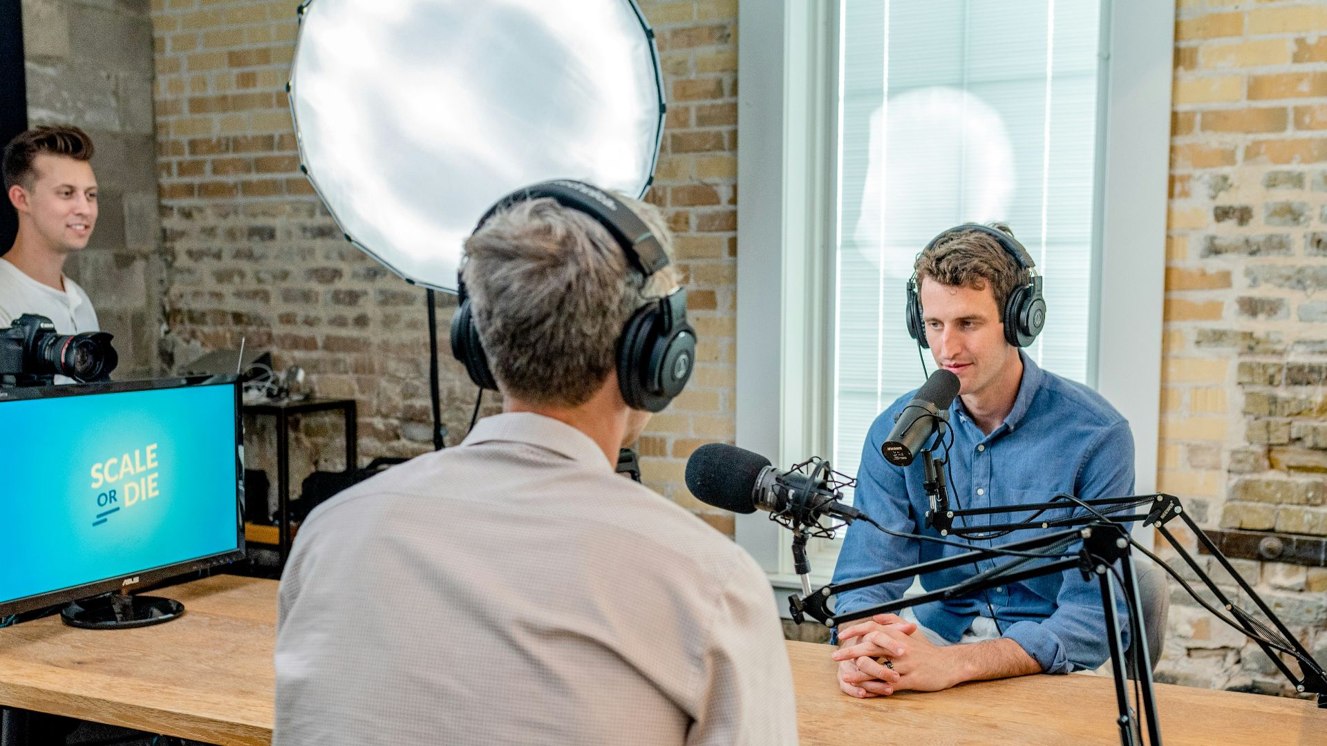 man in gray shirt leaning on table with headphones facing another man leaning on table with headboard