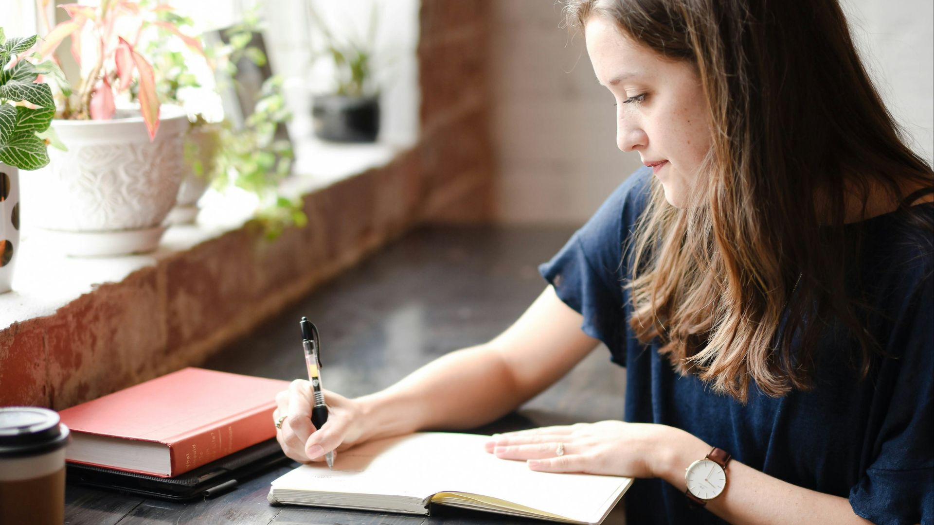 woman sitting in front of black table writing on white book near window