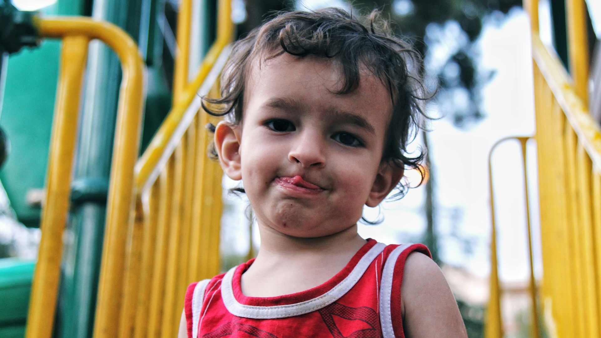 girl in red tank top standing near yellow metal fence during daytime
