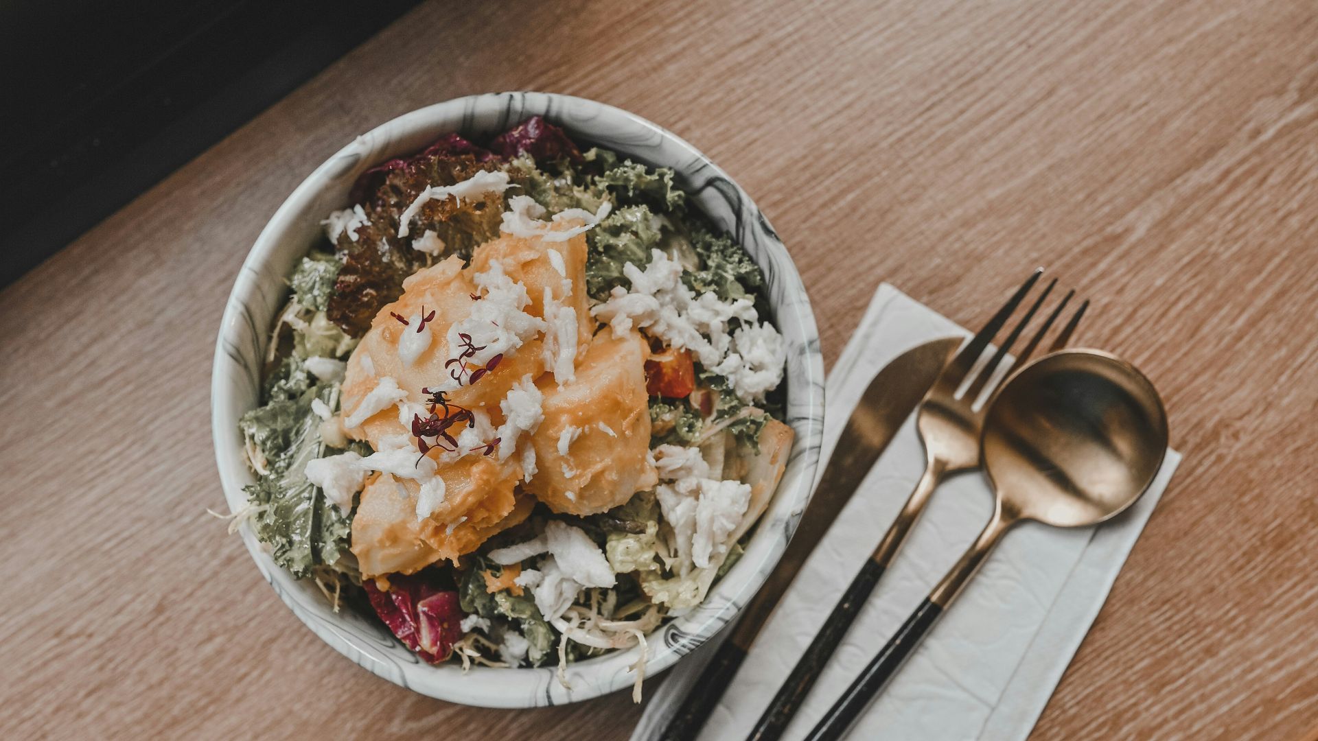 vegetable salad in white ceramic bowl beside stainless steel fork and knife on brown wooden table