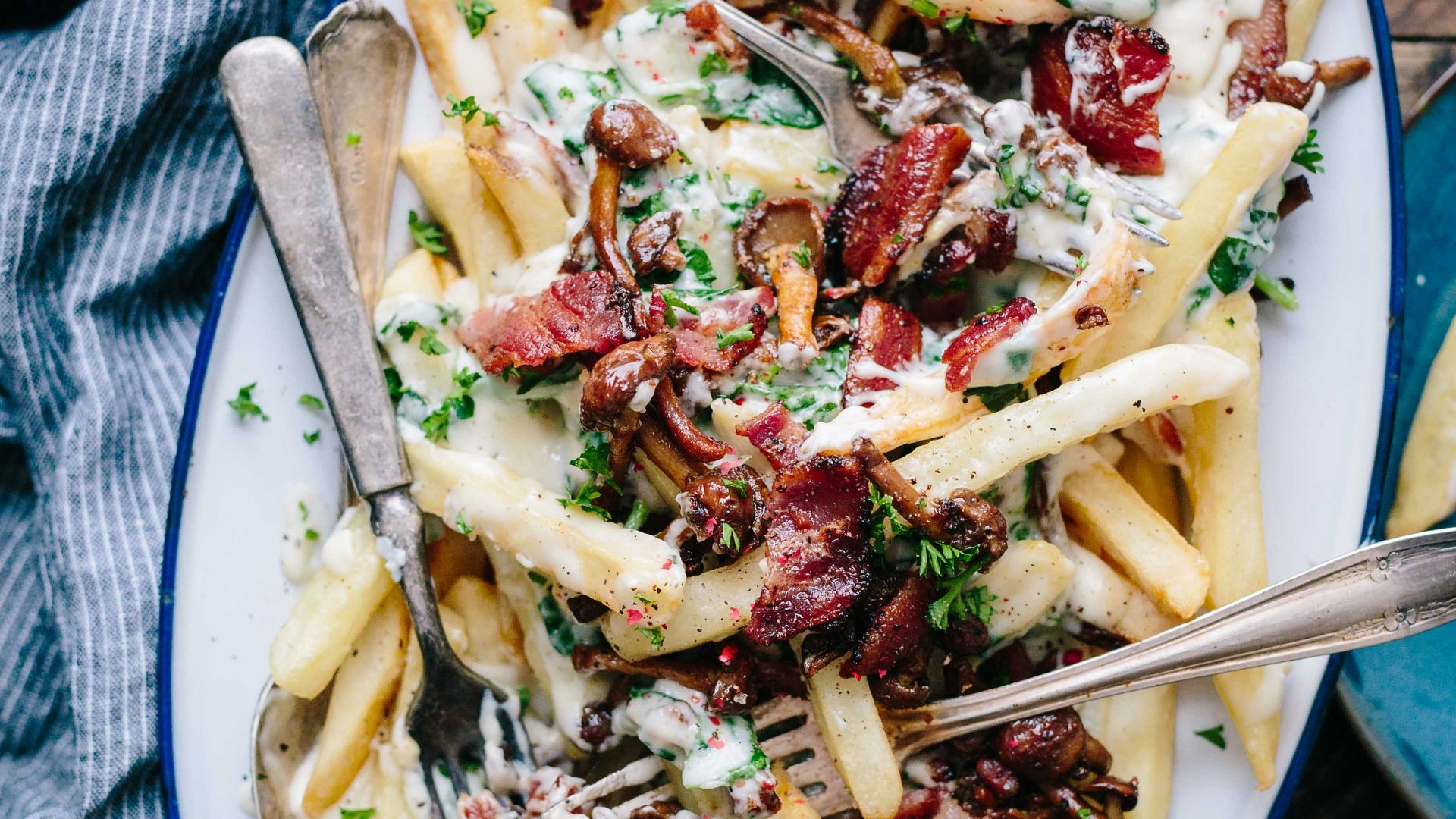bacon strips and melted cheese topped fries on oval white and blue platter with gray stainless steel forks