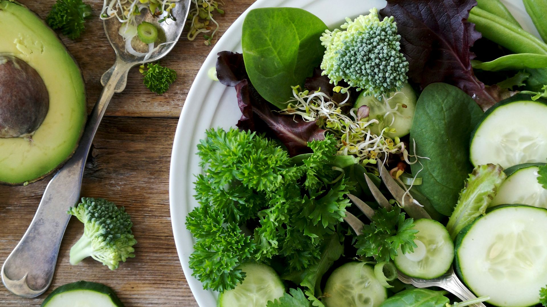 sliced broccoli and cucumber on plate with gray stainless steel fork near green bell pepper, snowpea, and avocado fruit