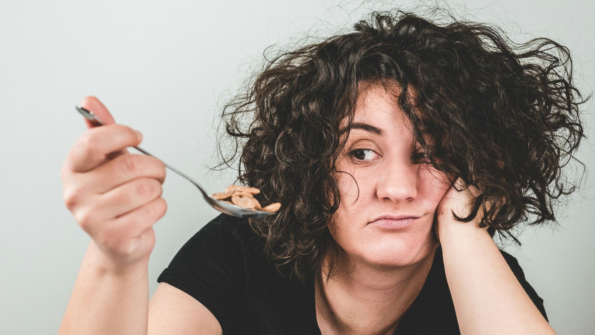 woman with messy hair wearing black crew-neck t-shirt holding spoon with cereals on top