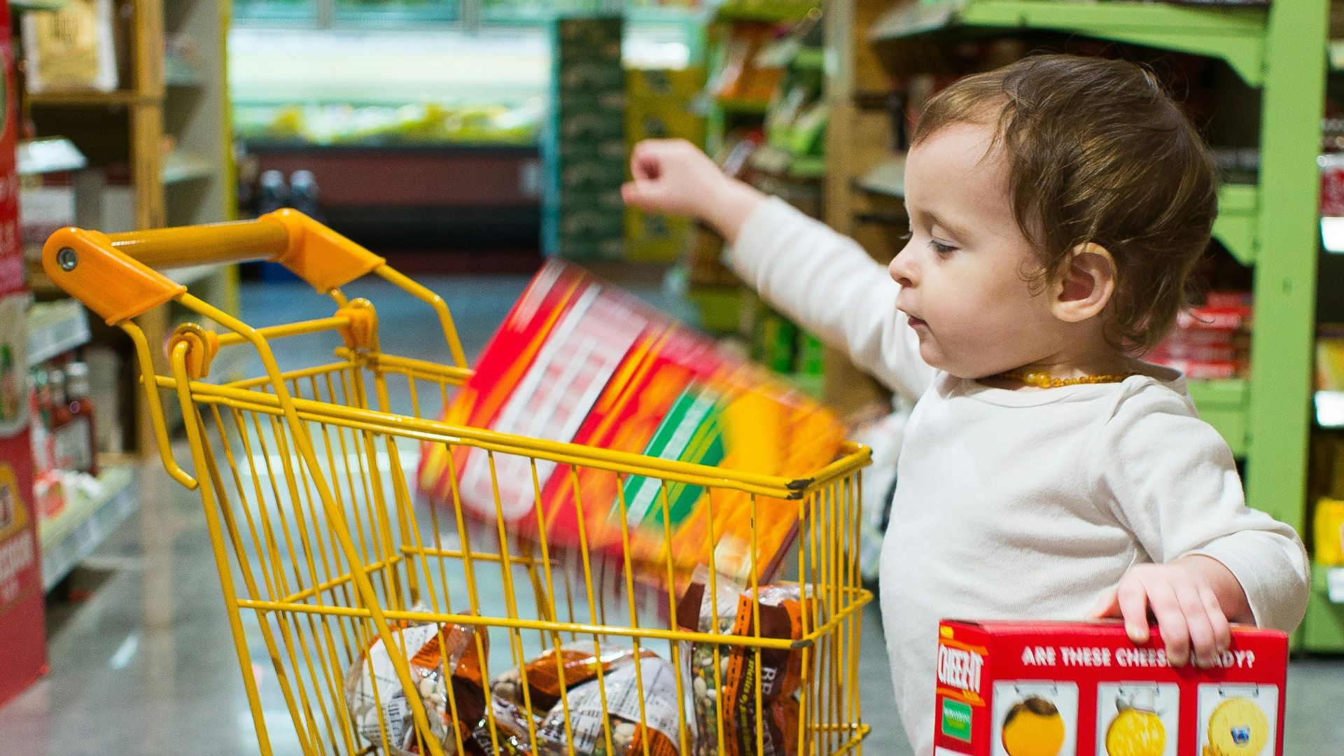 toddle carrying red and white box standing beside yellow shopping cart
