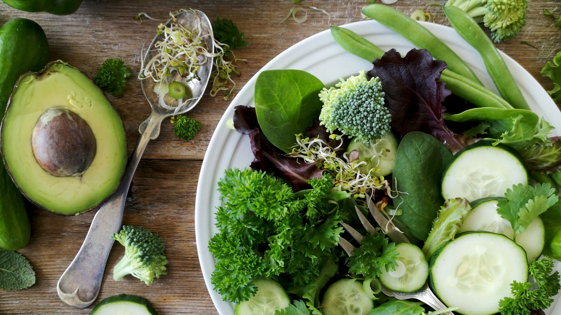 sliced broccoli and cucumber on plate with gray stainless steel fork near green bell pepper, snowpea, and avocado fruit