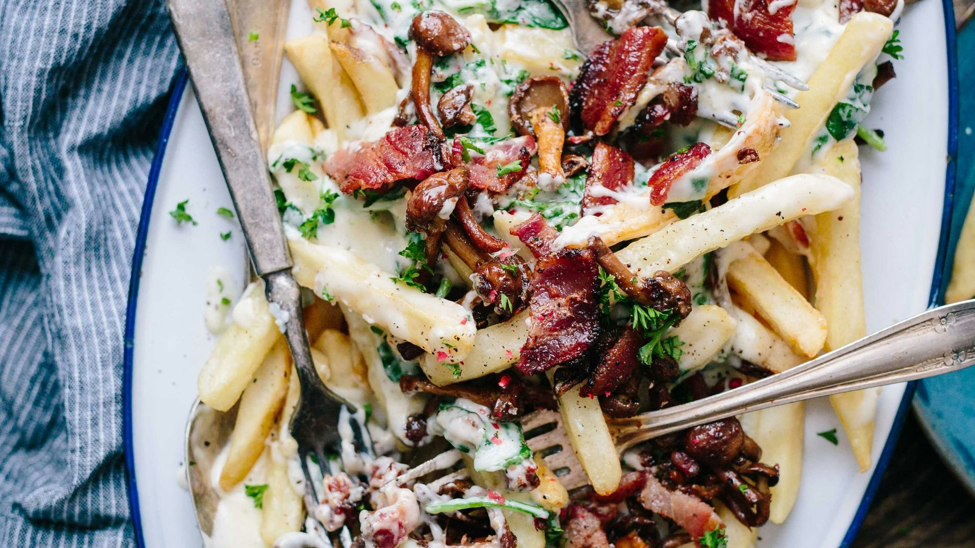 bacon strips and melted cheese topped fries on oval white and blue platter with gray stainless steel forks