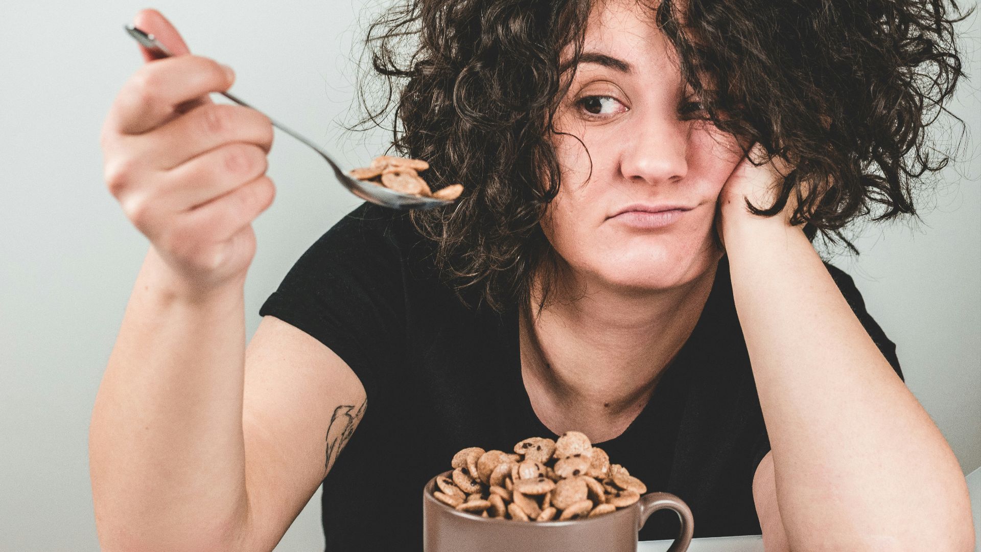 woman with messy hair wearing black crew-neck t-shirt holding spoon with cereals on top